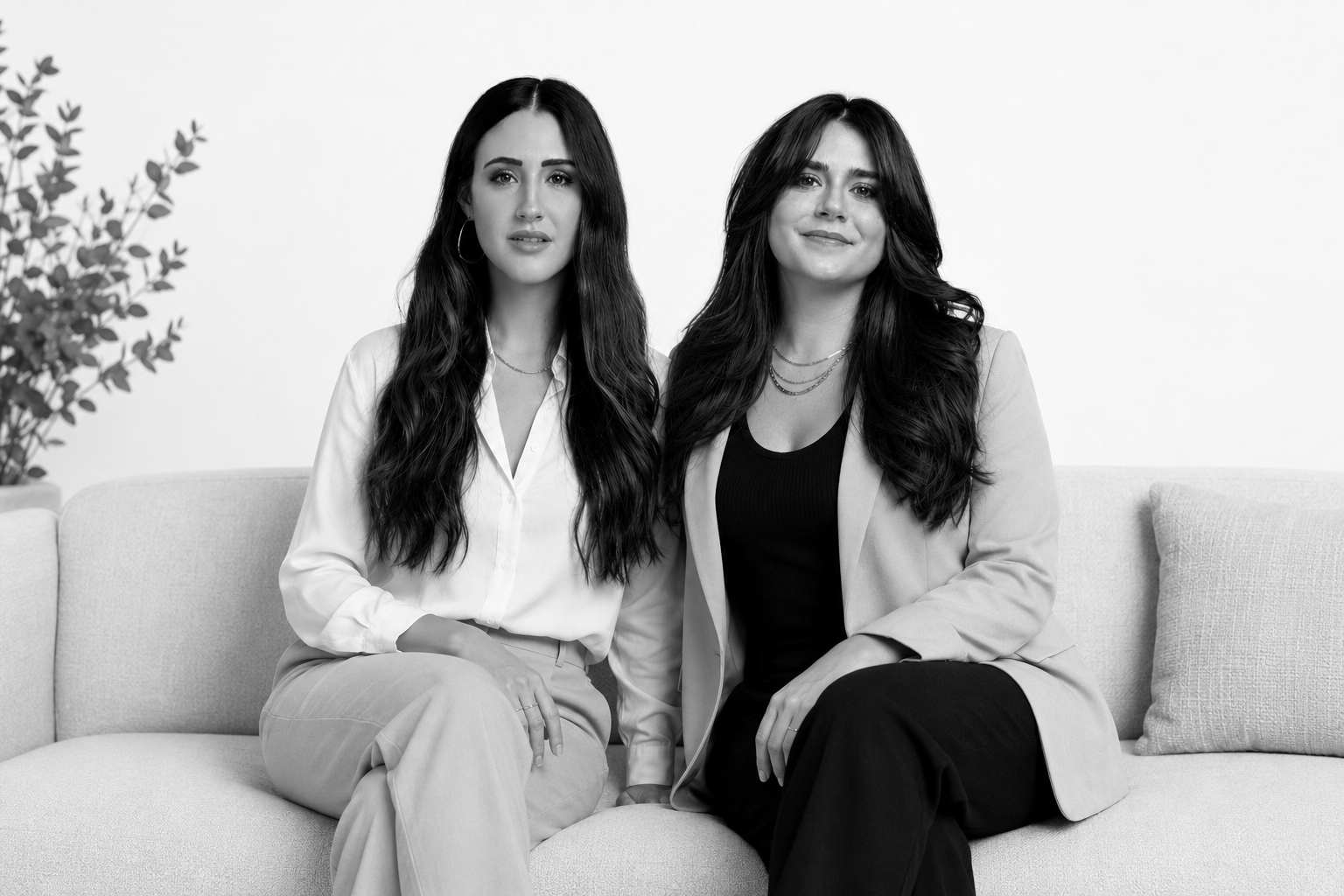 Two women with long dark hair sitting on a light-colored sofa in front of a plain white wall, one wearing a white blouse and the other wearing a blazer and a black top.