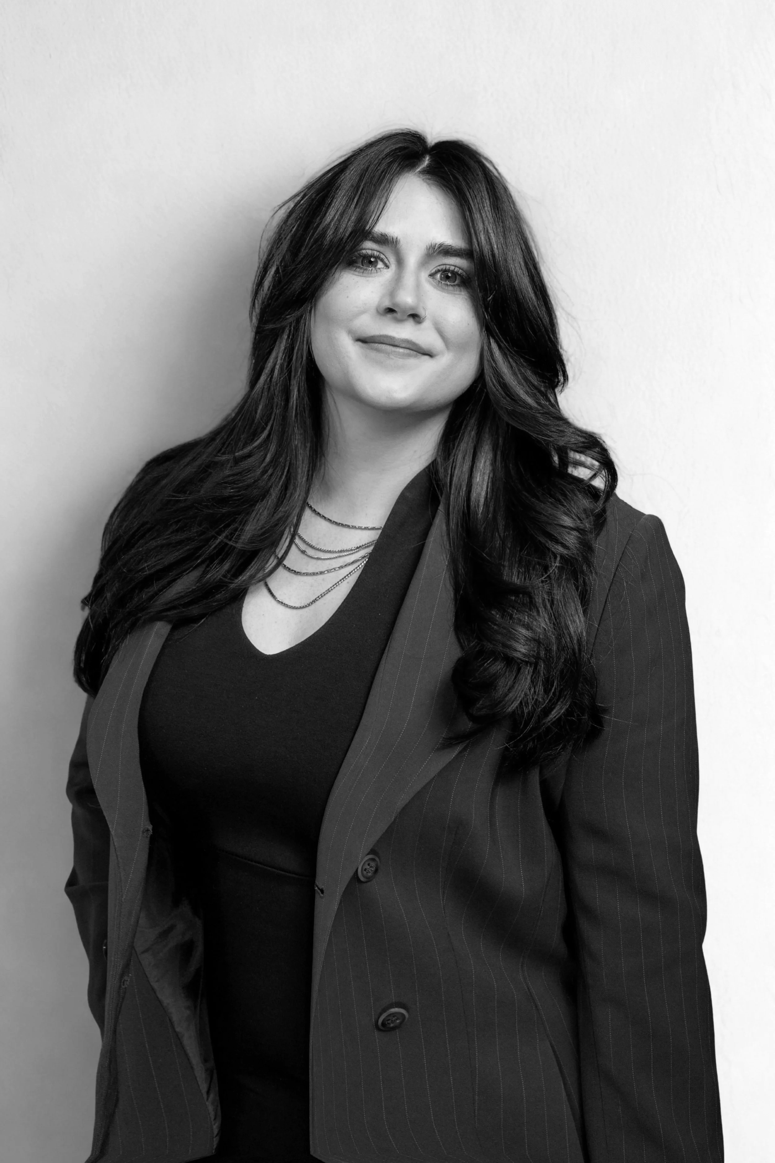 Black and white portrait of Rachel Reid with long wavy hair, wearing a pinstripe blazer and layered necklaces, standing against a plain wall.