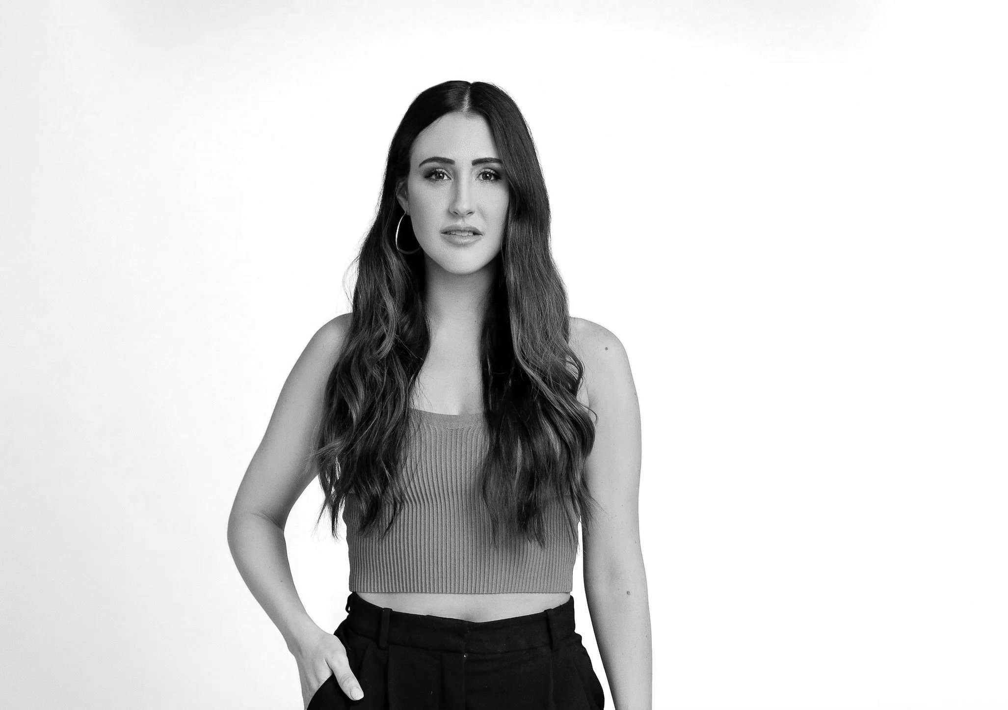 Black and white photo of Hannah Cox with long wavy hair, wearing hoop earrings, a sleeveless top, and dark pants, standing against a plain background.