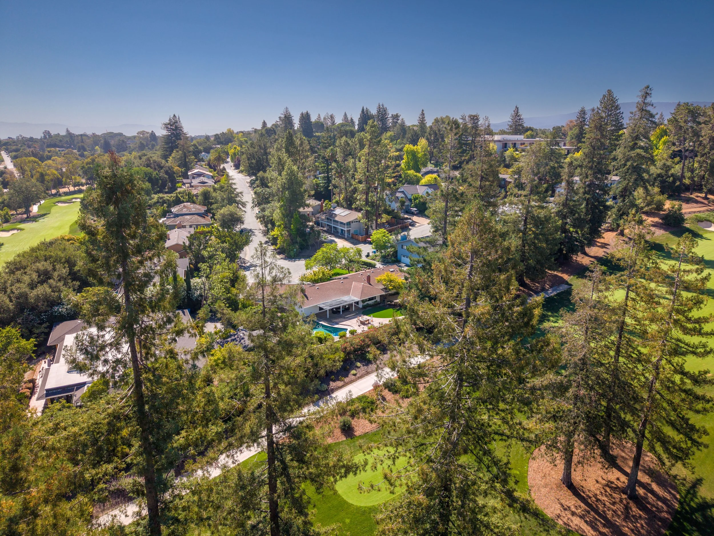 Aerial view of a suburban neighborhood with houses, a golf course, numerous trees, and a clear blue sky.