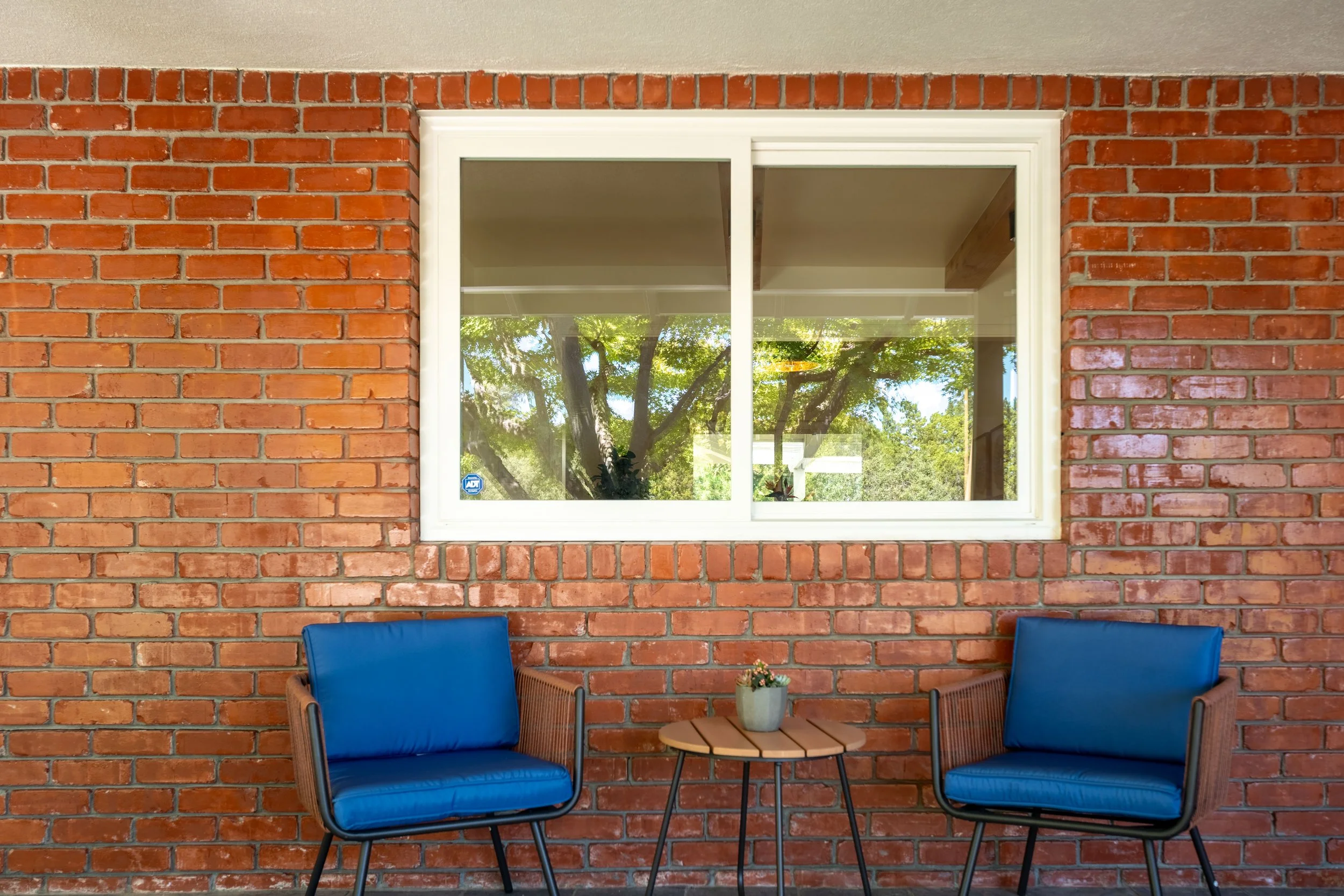A brick wall with a window reflecting trees, and two blue chairs with a small table and potted plant in front.