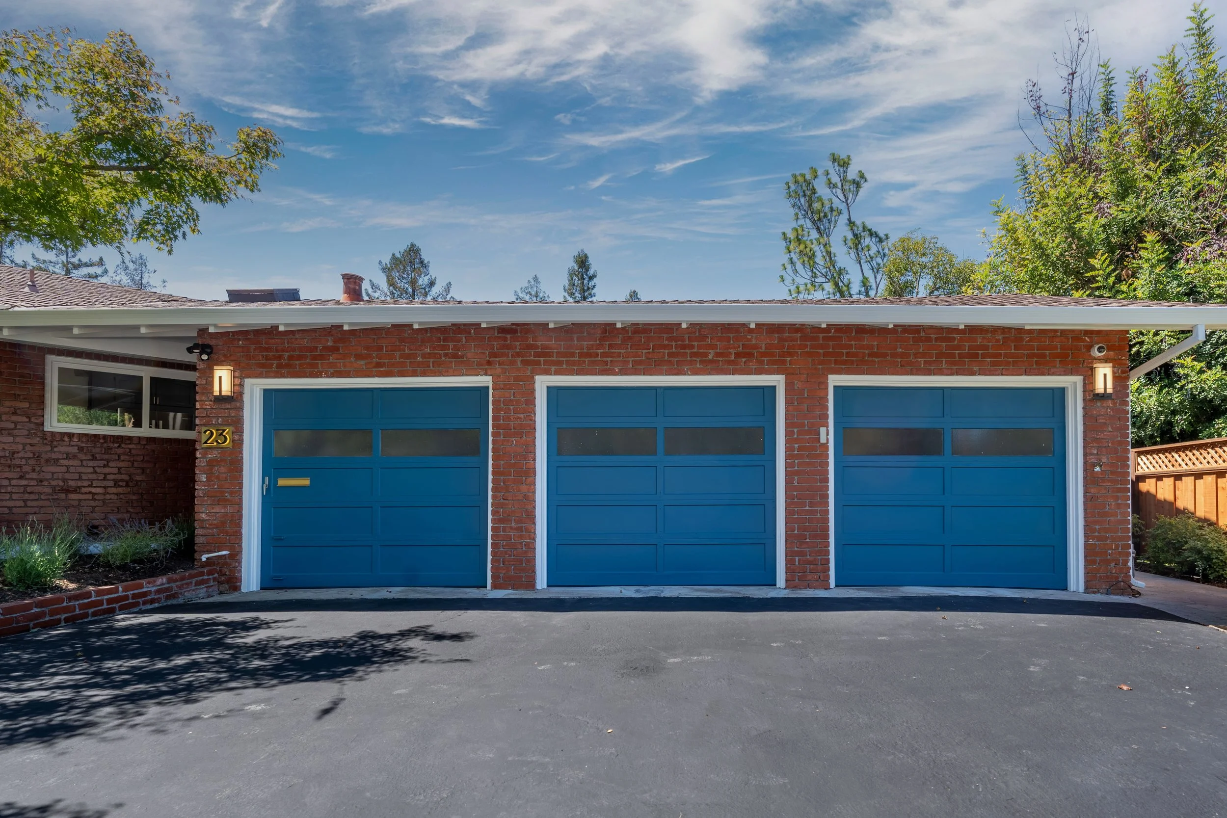 A red brick house with two blue garage doors, a small window to the left, and house number 23. The house has a white trim and roof, with a small landscaped garden area in front. The sky above is partly cloudy with green trees surrounding the house.
