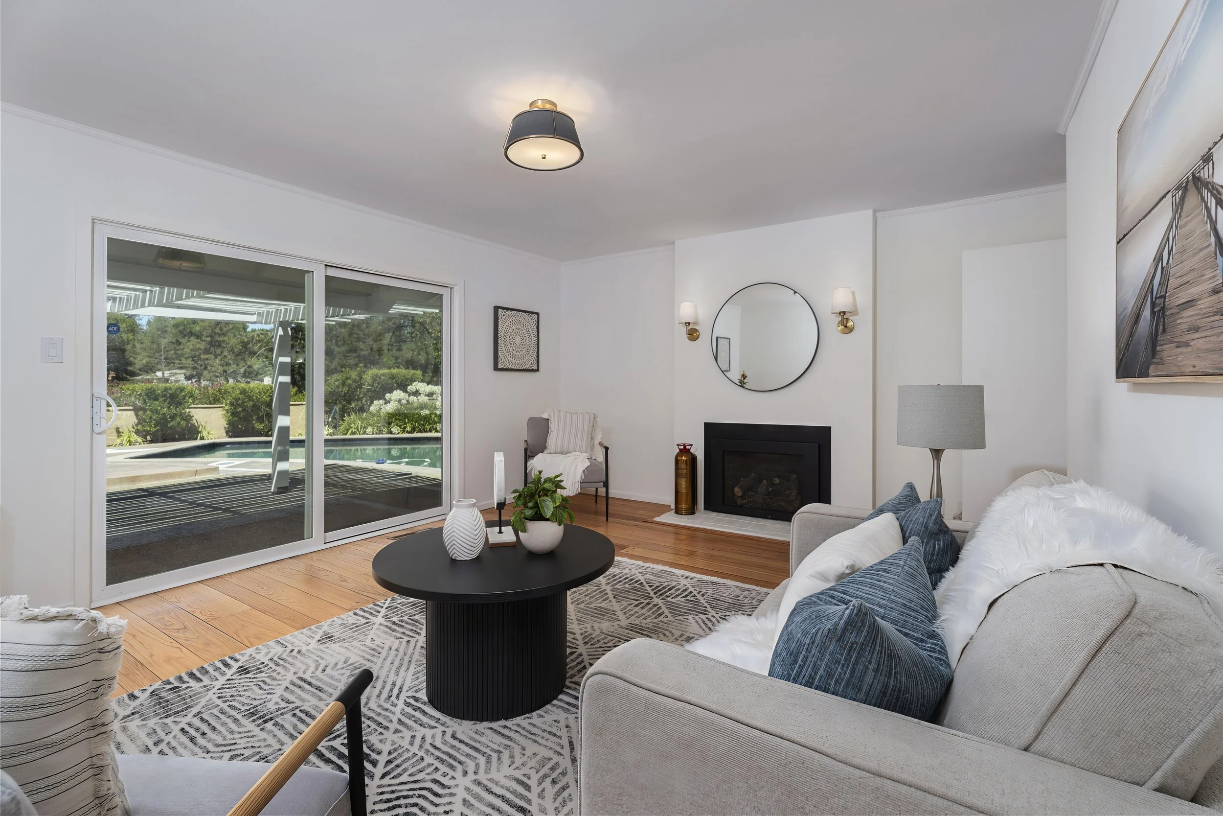 Living room with coffee table, beige sofa, throw pillows, white furry blanket, fireplace, wall mirror, wall sconces, artwork, sliding glass door, view of backyard with pool and trees.
