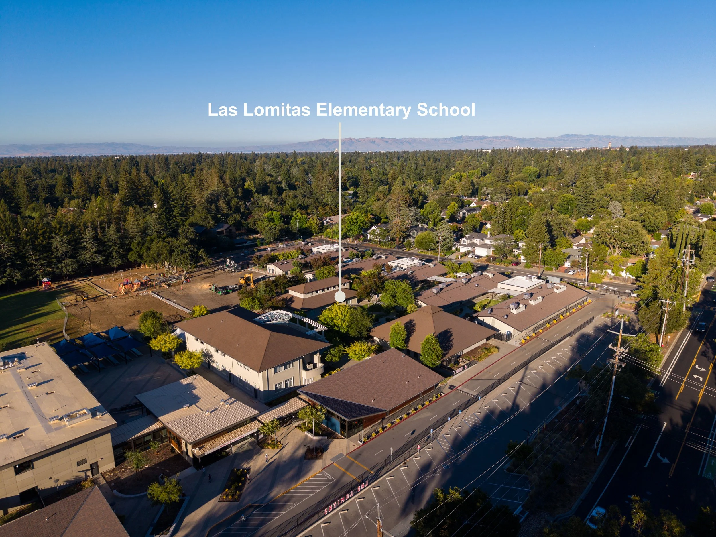 Aerial view of a neighborhood with houses, trees, and streets, with mountains in the background under a clear blue sky. The text overlay reads 'Las Lomitas Elementary School'.