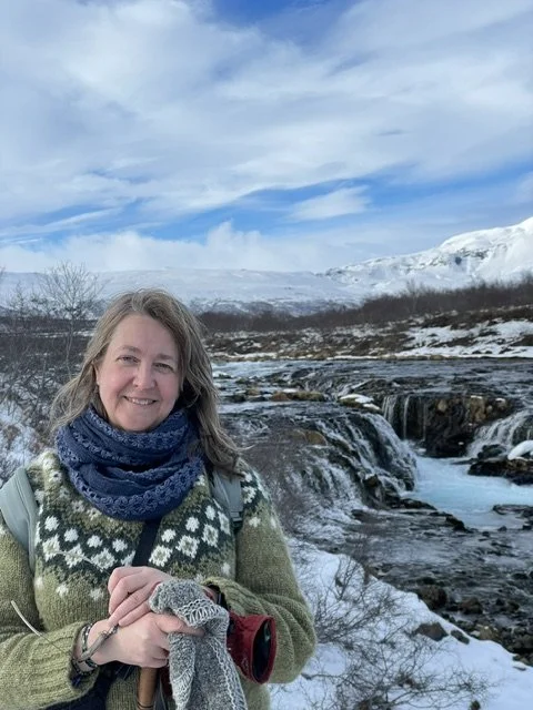 Michele standing outdoors next to a river with waterfalls, snow-covered hills, and a partly cloudy sky in the background.