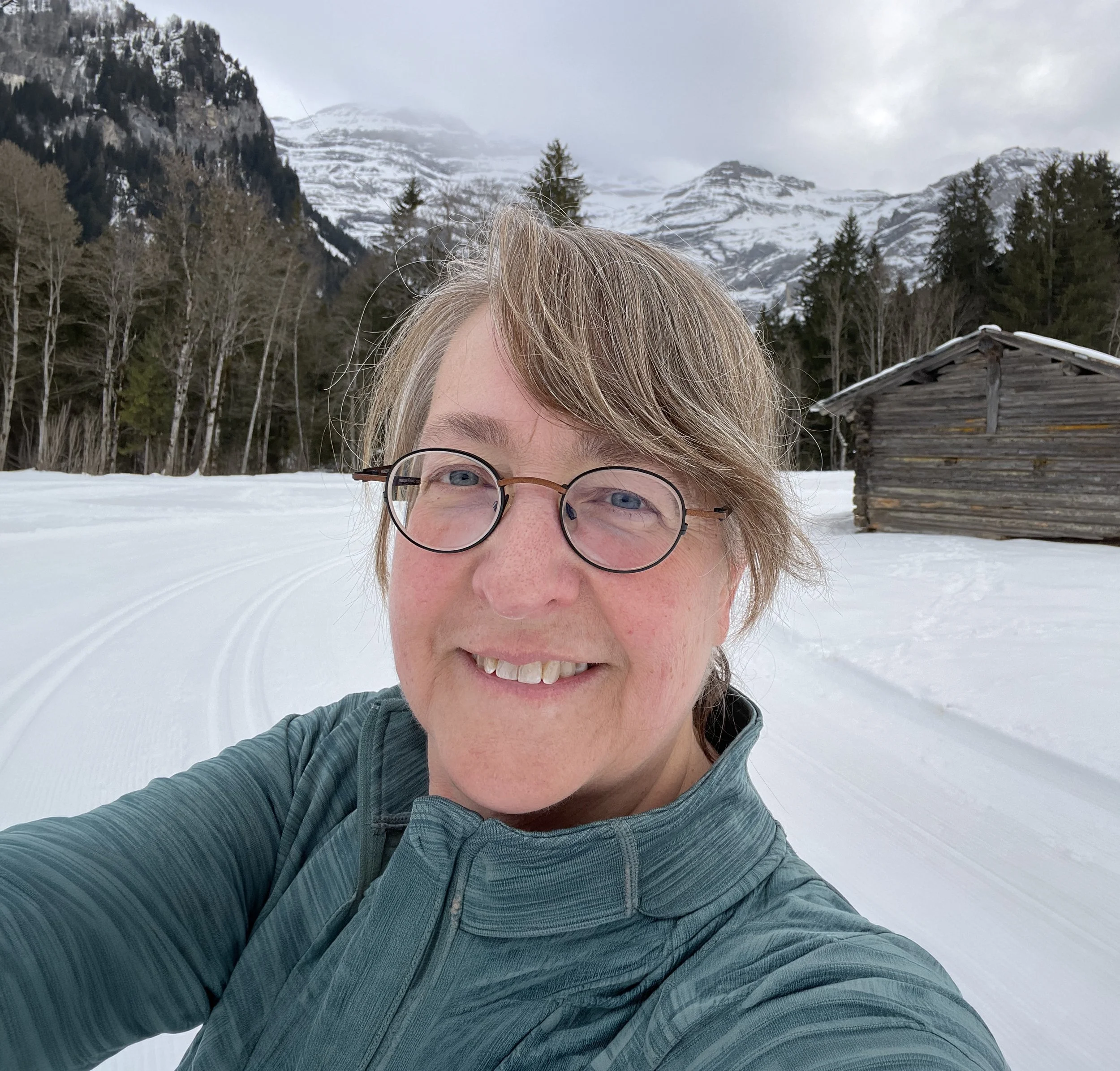 Michele taking a selfie outdoors in a snowy mountain landscape with trees, a wooden cabin, and snow-covered peaks in the background.