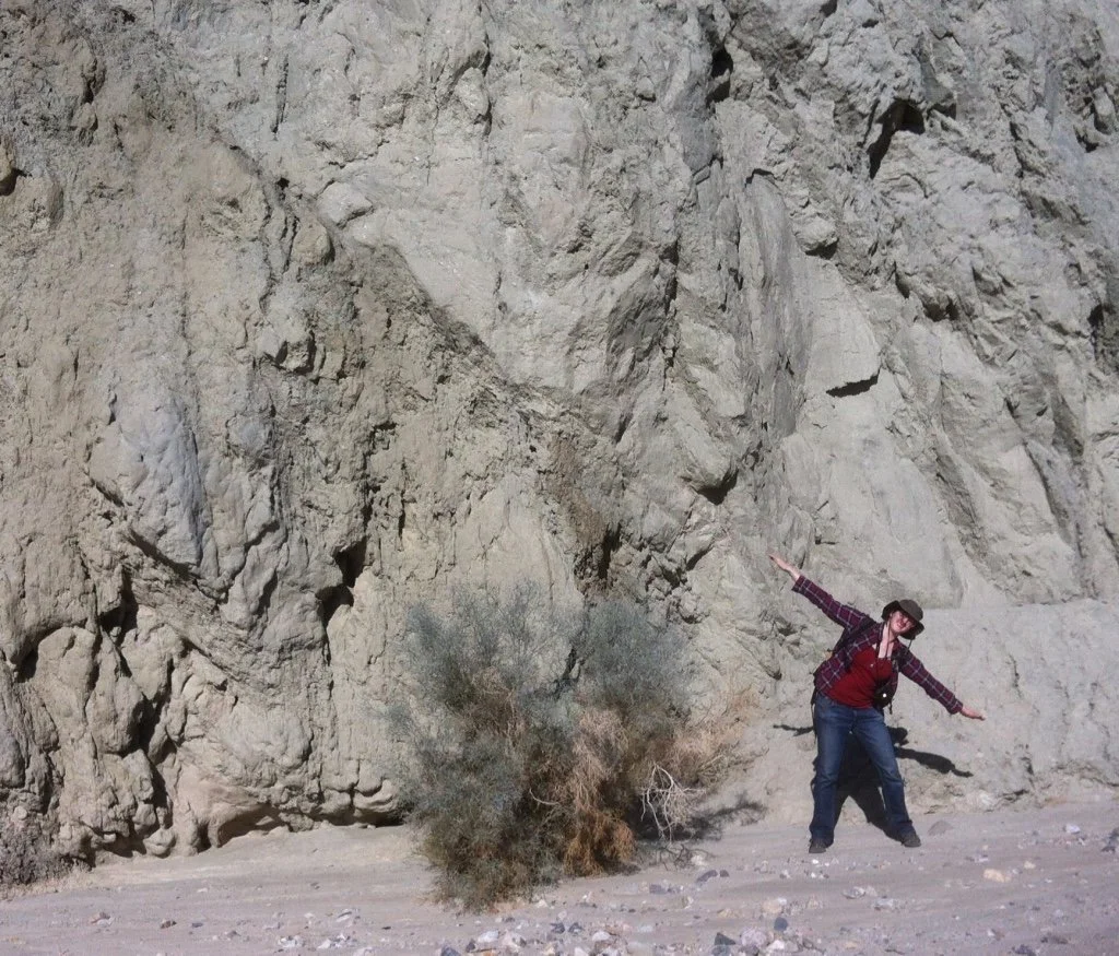Person with a hat and plaid shirt standing in a desert, arms outstretched as the same fault line in front of a large rocky wall.