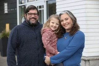 A family of three posing outdoors in front of a house, smiling at the camera. The father has glasses and a beard, the mother has long hair, and their young daughter is being held by the mother.
