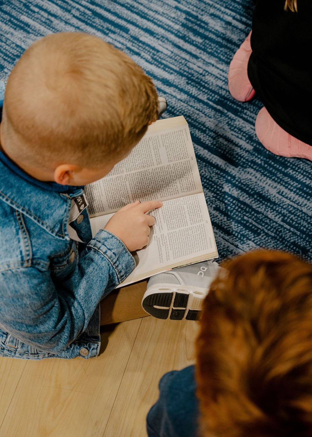 A young boy with blonde hair wearing a denim jacket sitting on the floor, reading a book and pointing at the pages. Another person with red hair is also sitting nearby, and a girl in pink socks is partially visible kneeling next to him.