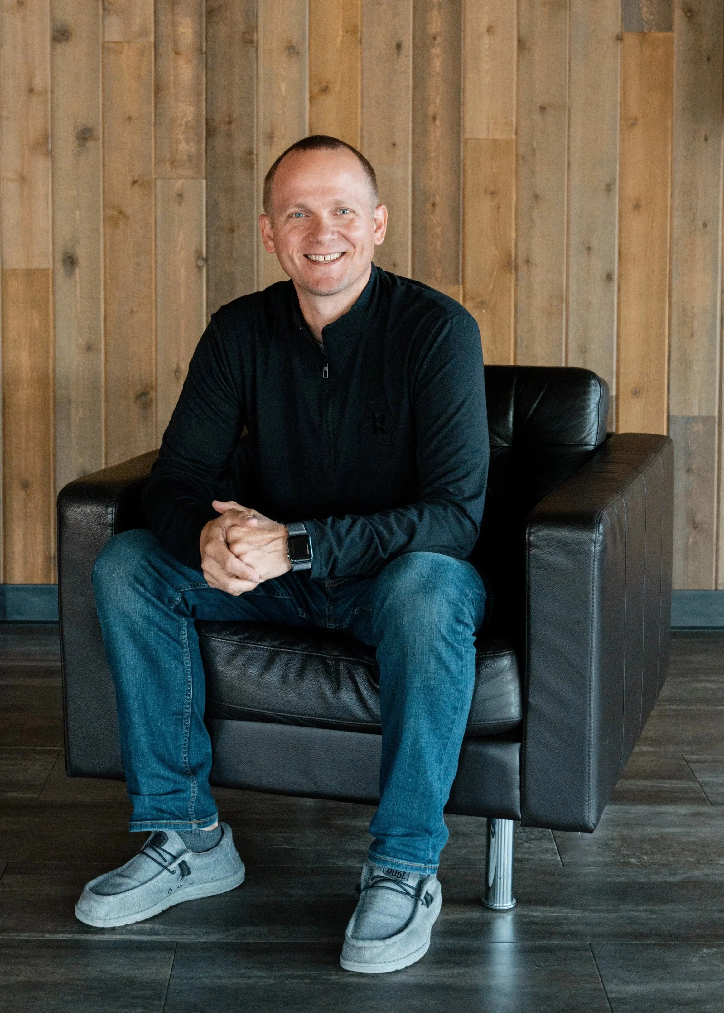 A man sitting on a black leather chair in front of a wooden wall, smiling, wearing a black zip-up jacket, blue jeans, gray sneakers, and a smartwatch. This is Landon Yowell, The Refinery Church's Lead Pastor.