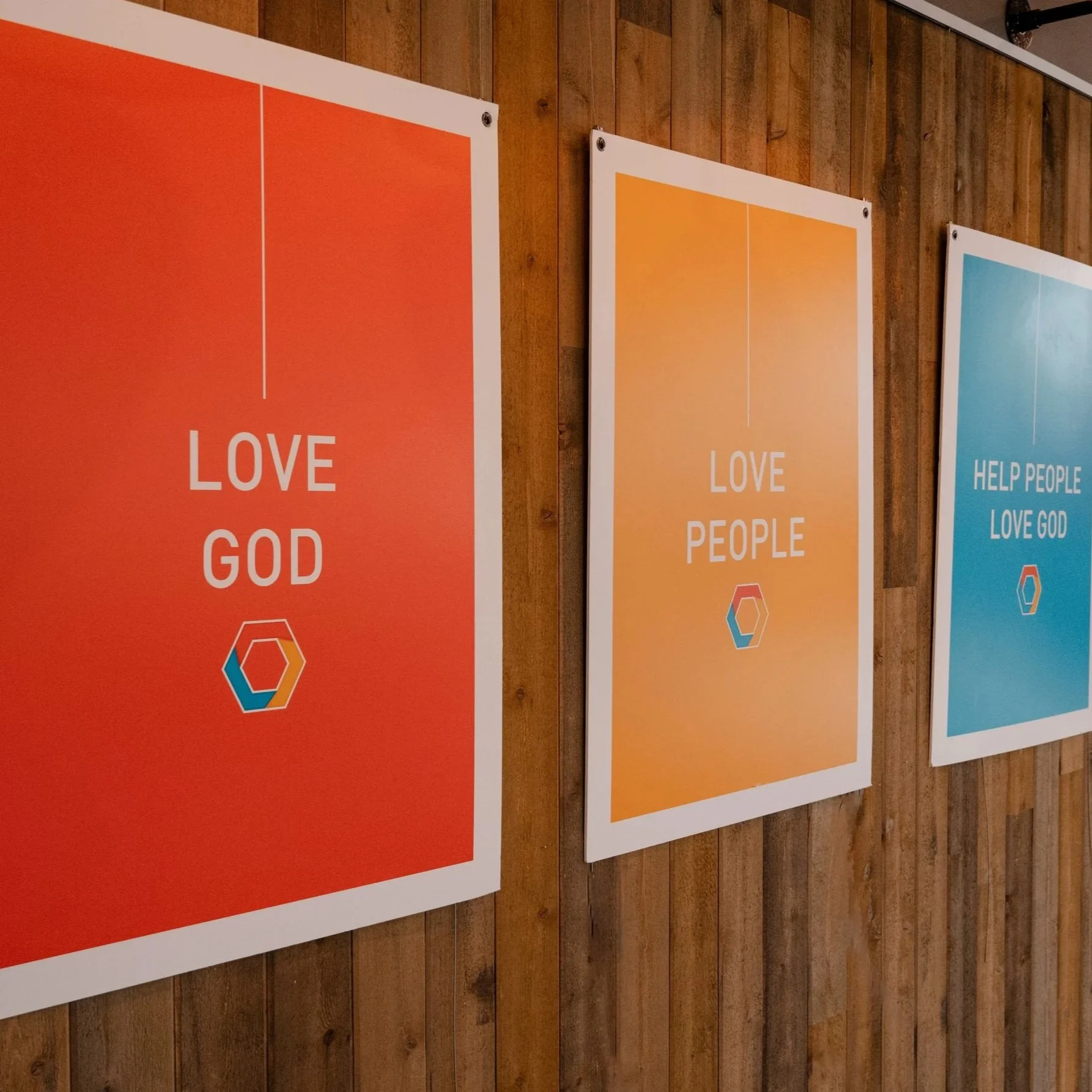 Three posters hanging on a wooden wall. The first poster is orange and says "LOVE GOD." The second poster is yellow and says "LOVE PEOPLE." The third poster is blue and says "HELP PEOPLE LOVE GOD."