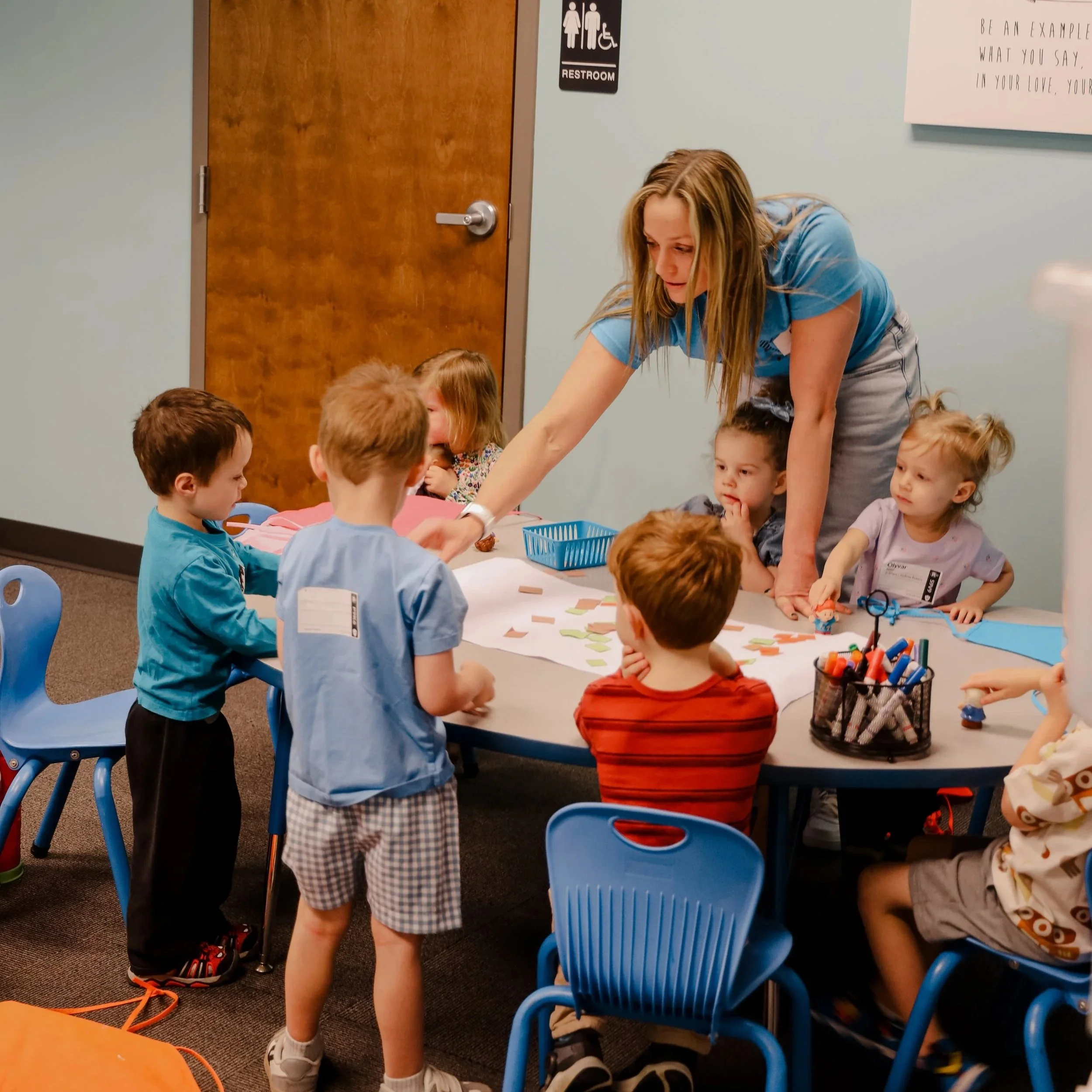 A group of young children seated at a table in a classroom, engaging in a craft or activity, with an adult assisting. The children are wearing colorful clothing and the classroom has blue chairs and a wall with a door and restroom sign.