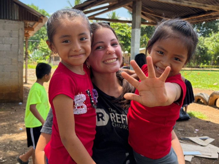 A woman smiling and holding two young girls in red shirts outdoors under a shelter, with a girl reaching out towards the camera. Background shows trees, a boy in a bright green shirt, and a grassy area.