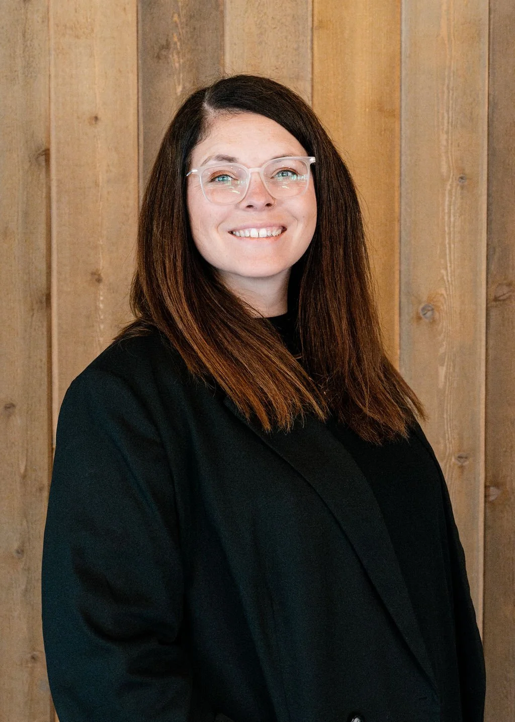 A woman with long brown hair, glasses, and a black blazer smiling in front of a wooden wall.