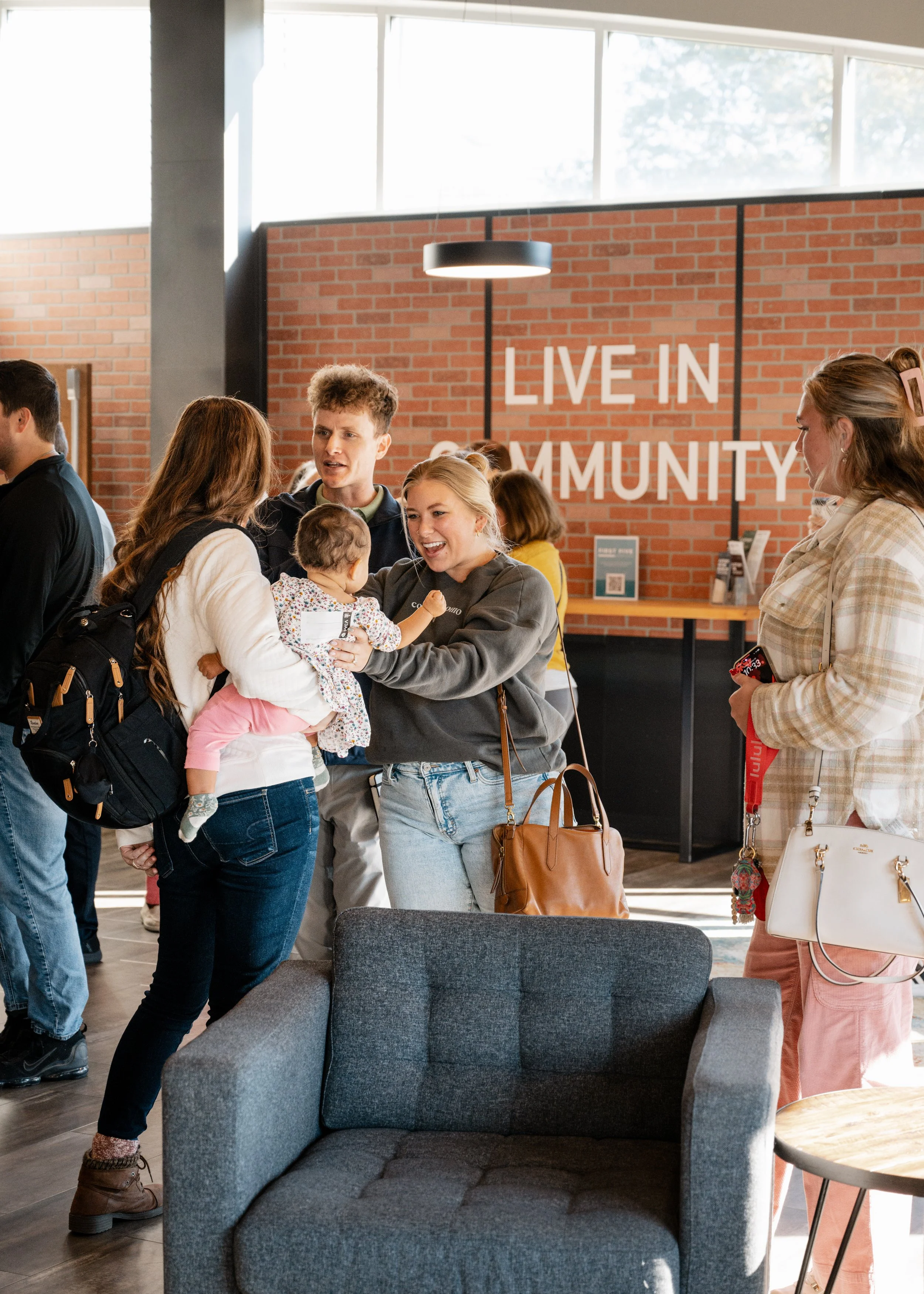 People socializing at a community event with a brick wall and large windows in the background, and a sign that reads 'LIVE IN COMMUNITY'.