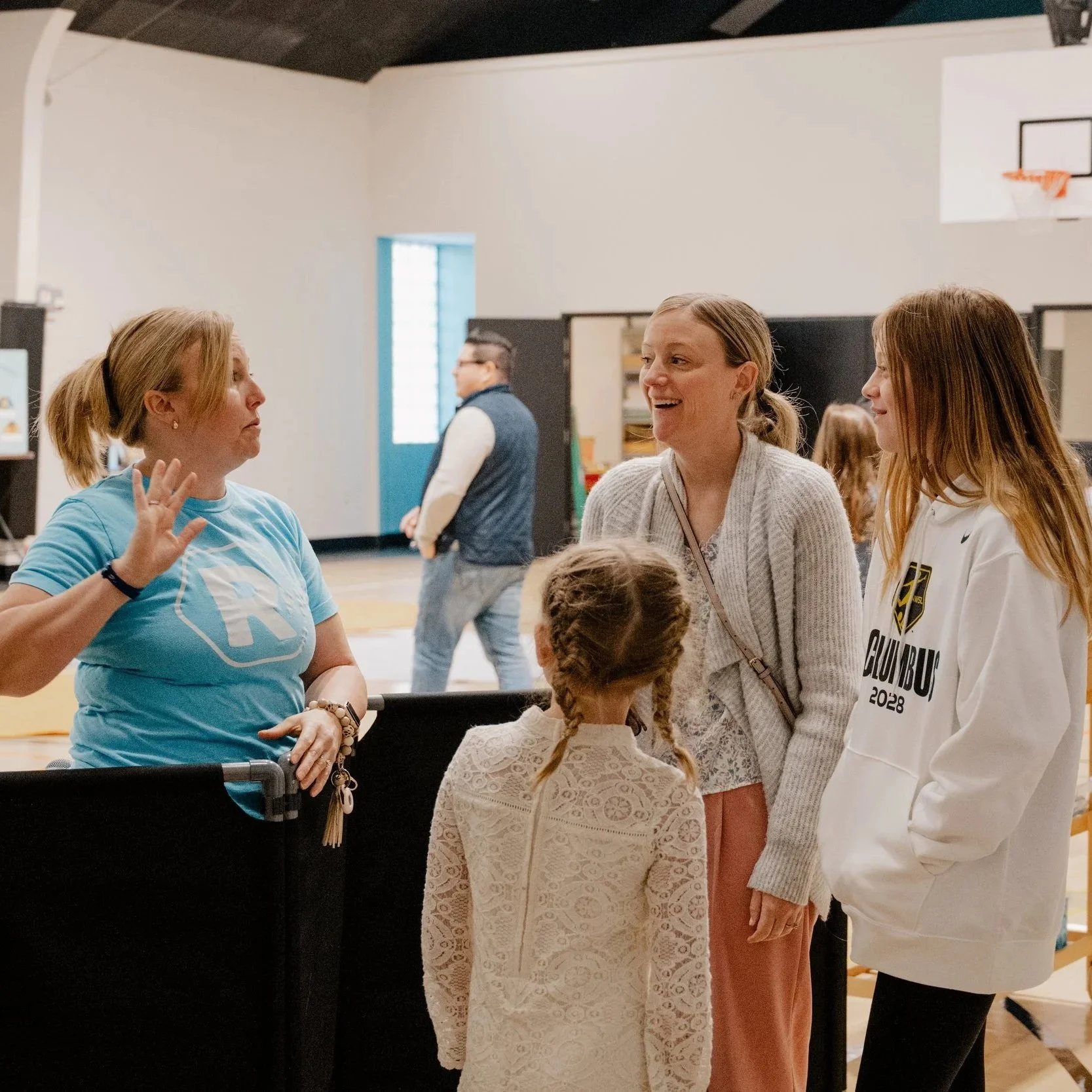 Group of people engaging in conversation in a gymnasium with a basketball hoop in the background.