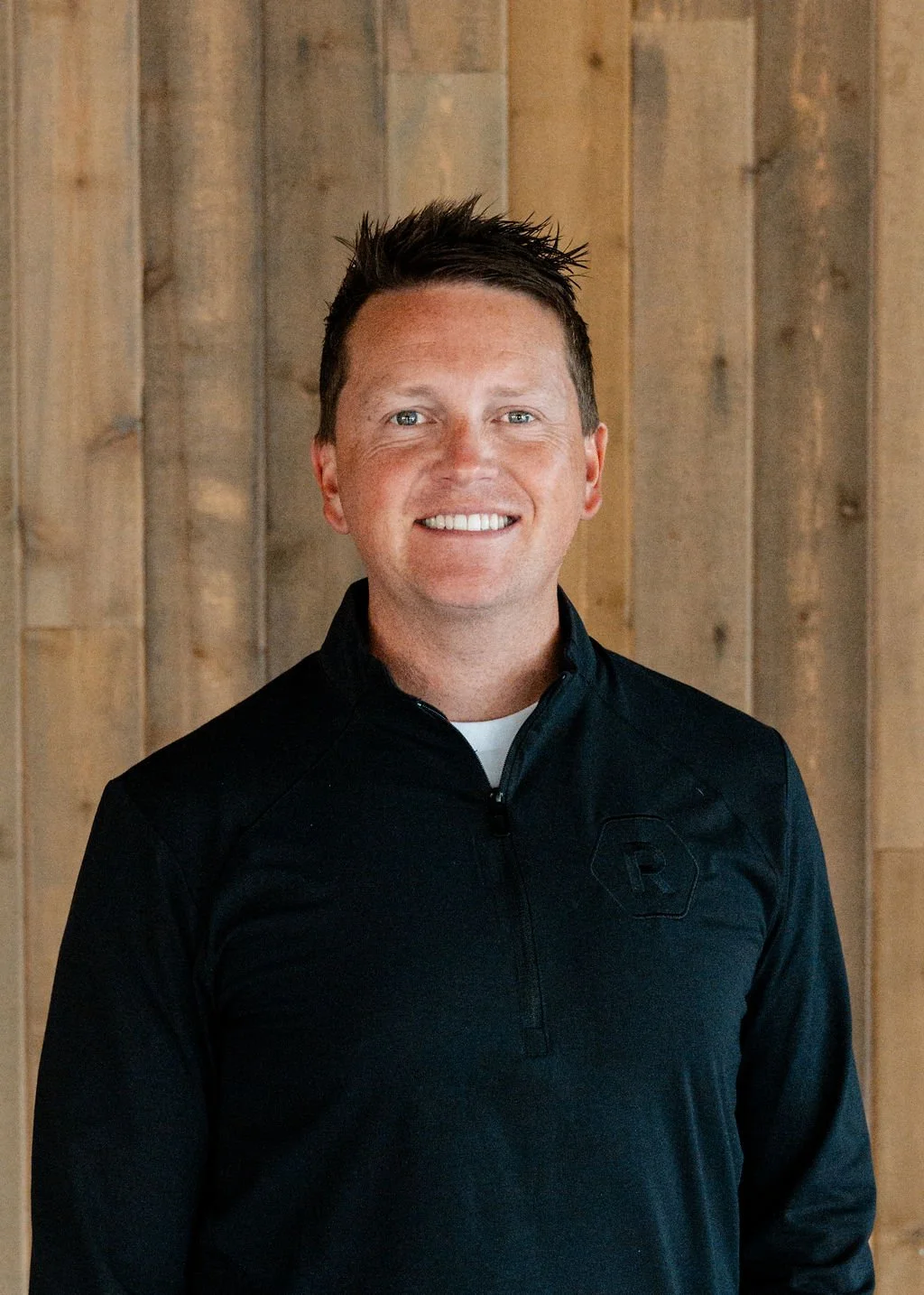 Smiling man with short dark hair in black zip-up jacket standing in front of a wooden wall.
