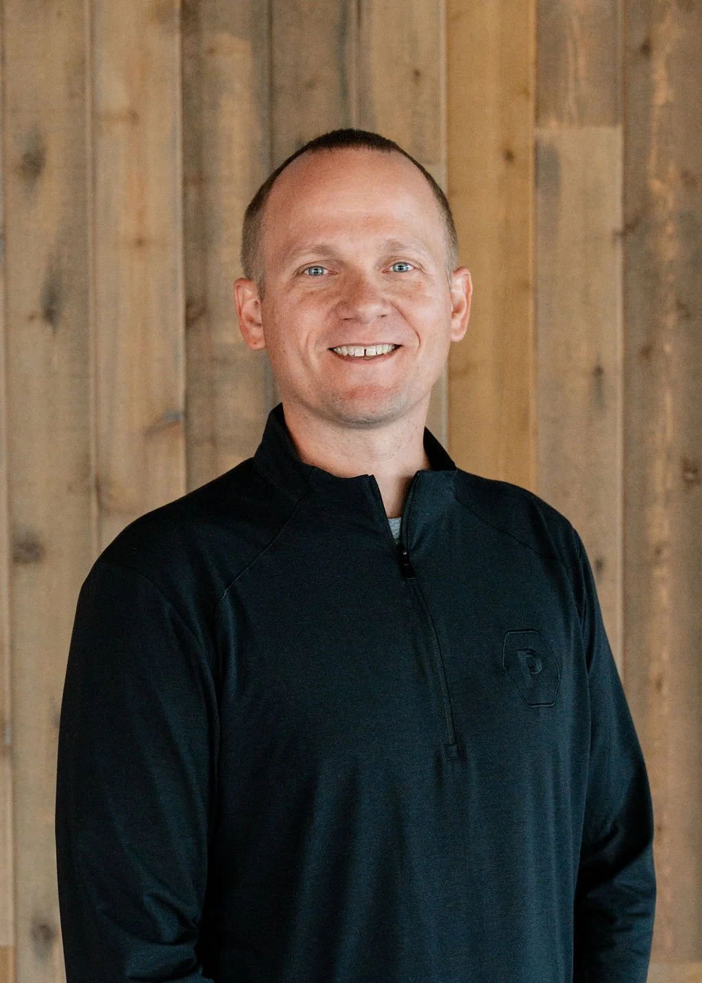 A smiling man with short light hair wearing a black quarter-zip sports jacket standing in front of a wooden plank wall.