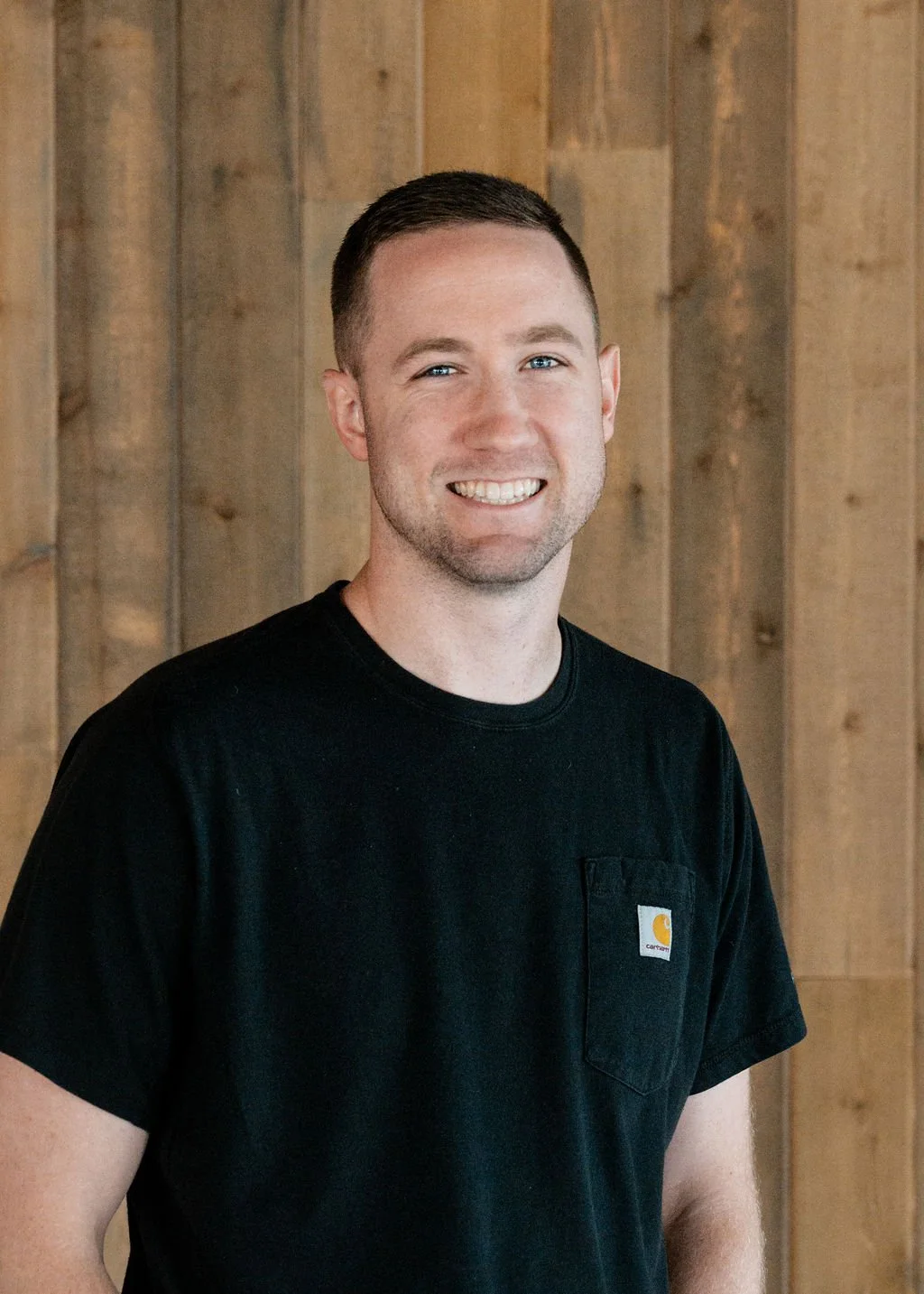 A smiling young man with short dark hair and a light beard, wearing a black Carhartt T-shirt, standing in front of a wooden wall.