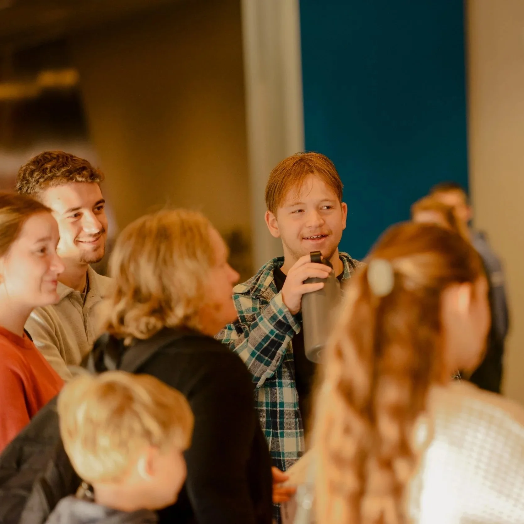 A group of people attending a presentation or conference, with a young man with red hair and a plaid shirt holding a water bottle among them.