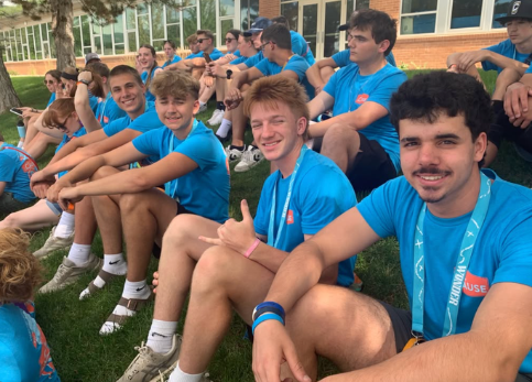 Group of teenage boys wearing matching blue shirts sitting on grass outside a building, smiling and giving thumbs-up signs during daytime.