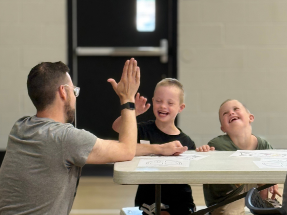A man with glasses giving a high five to a boy with short blonde hair, both smiling, while another boy with short blonde hair looks on and laughs, sitting at a table.