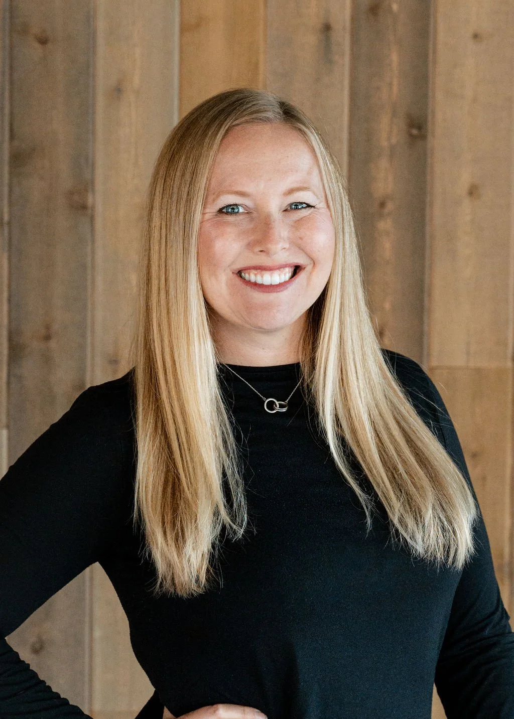 Portrait of a smiling woman with long blonde hair, wearing a black top and a silver necklace, standing against a wooden wall background.
