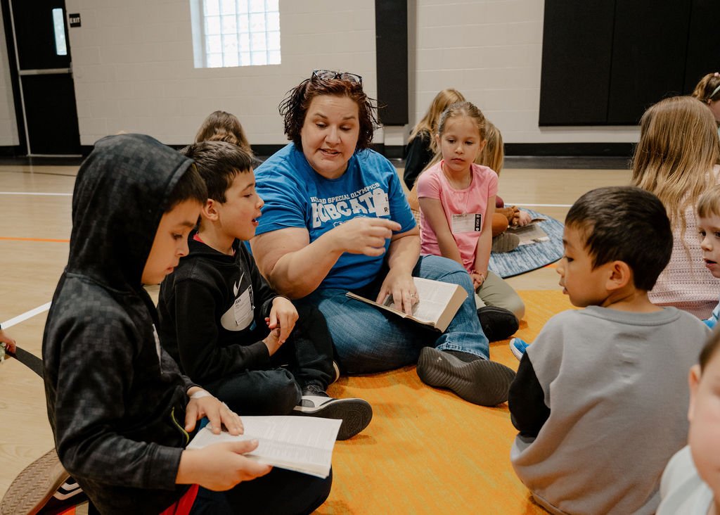 A woman sitting on the floor and reading a book to a group of young children in a gymnasium.