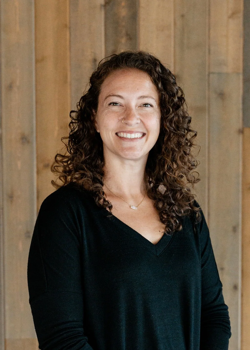 A smiling woman with curly hair, wearing a black top, standing in front of a wooden wall.