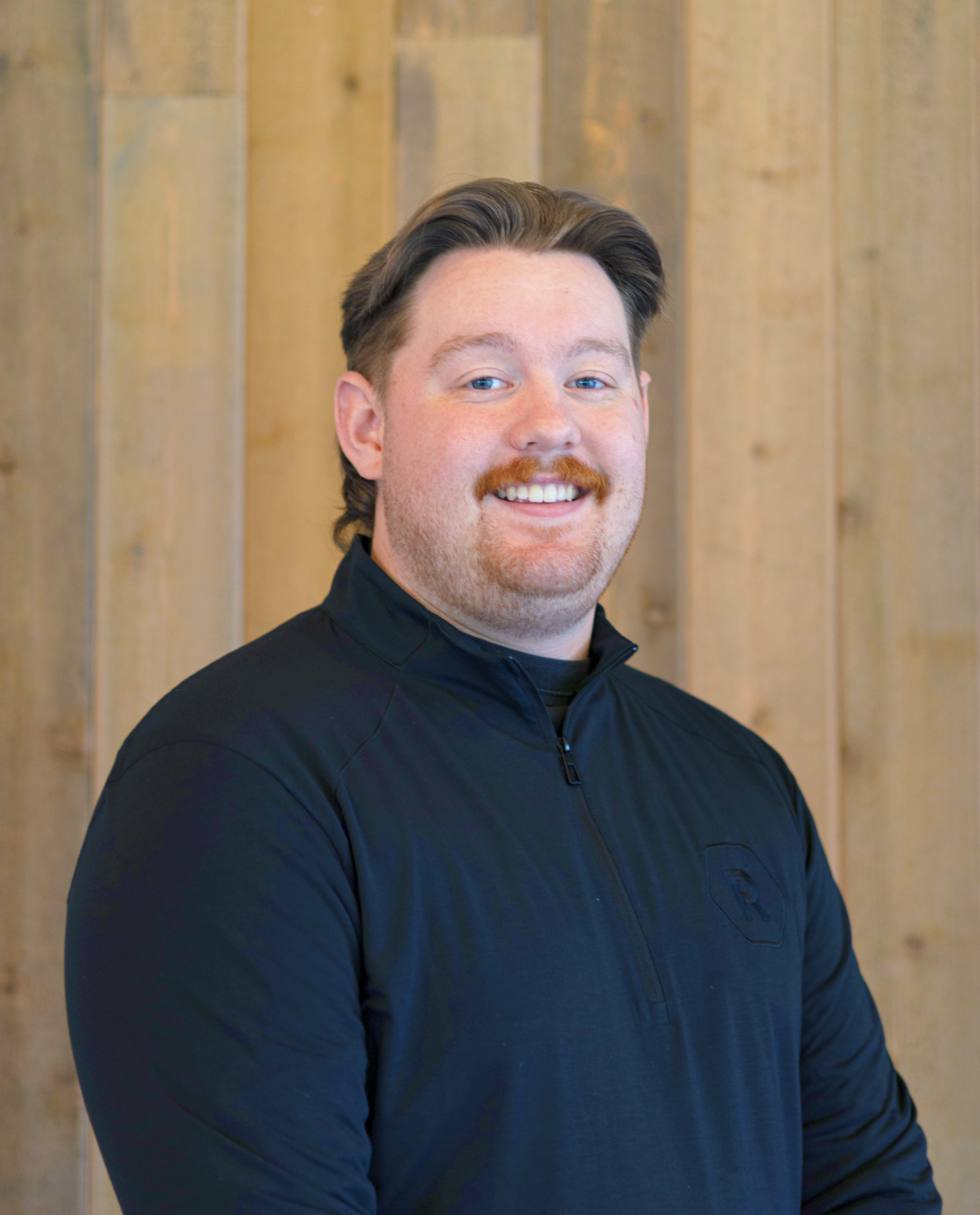 A smiling man with brown hair, a mustache, and a beard, wearing a black zip-up jacket, standing in front of a wooden wall.