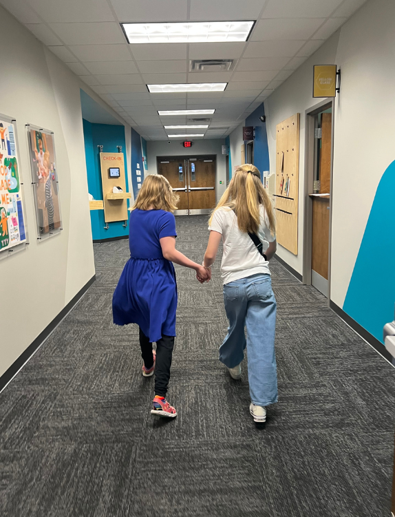 Two young girls holding hands walking down a school hallway with bulletin boards and doors.
