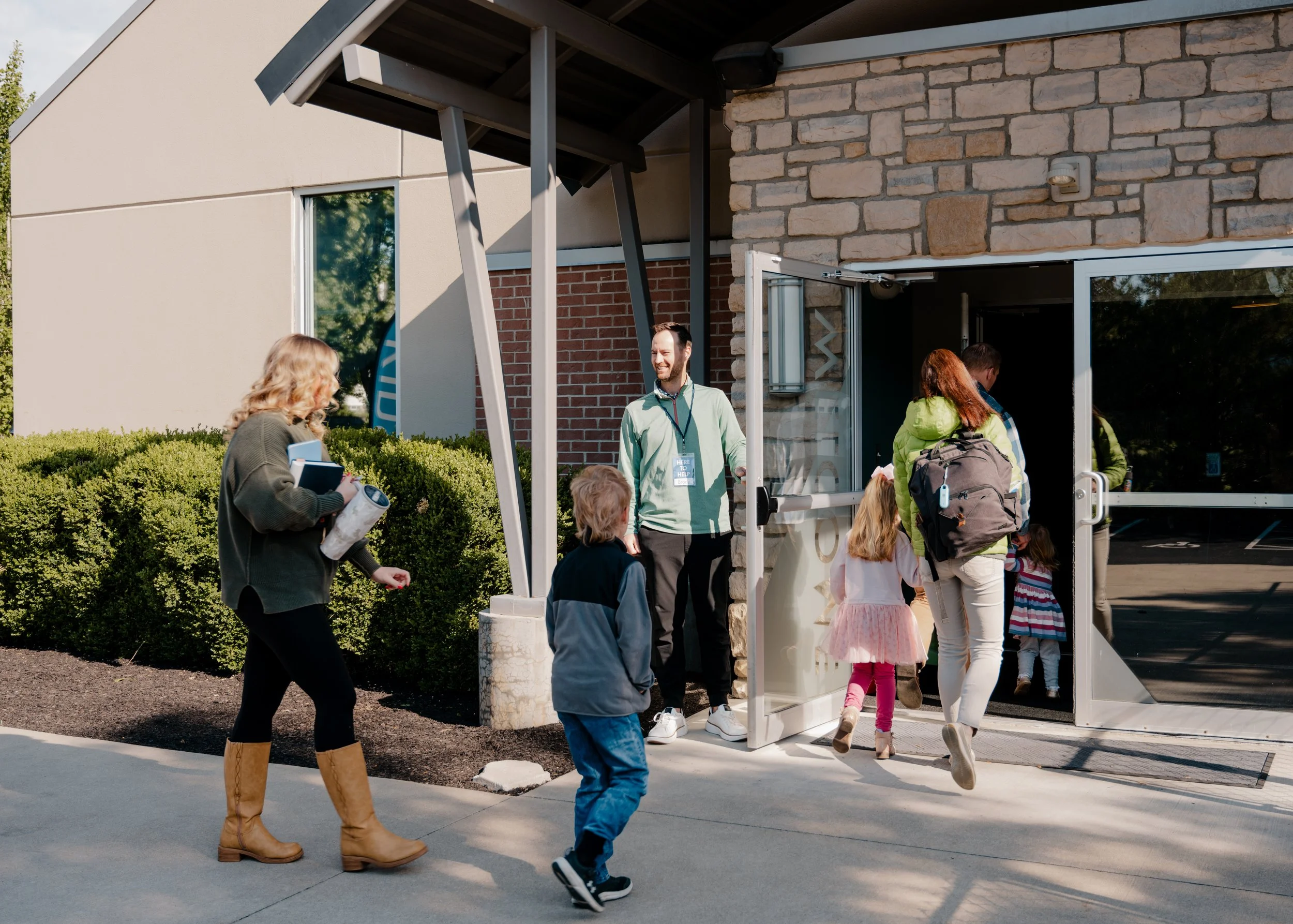 A group of people, including children and adults, walking out of a building with a glass door. The building has a brick and stucco exterior with greenery and bushes nearby. It is a sunny day.