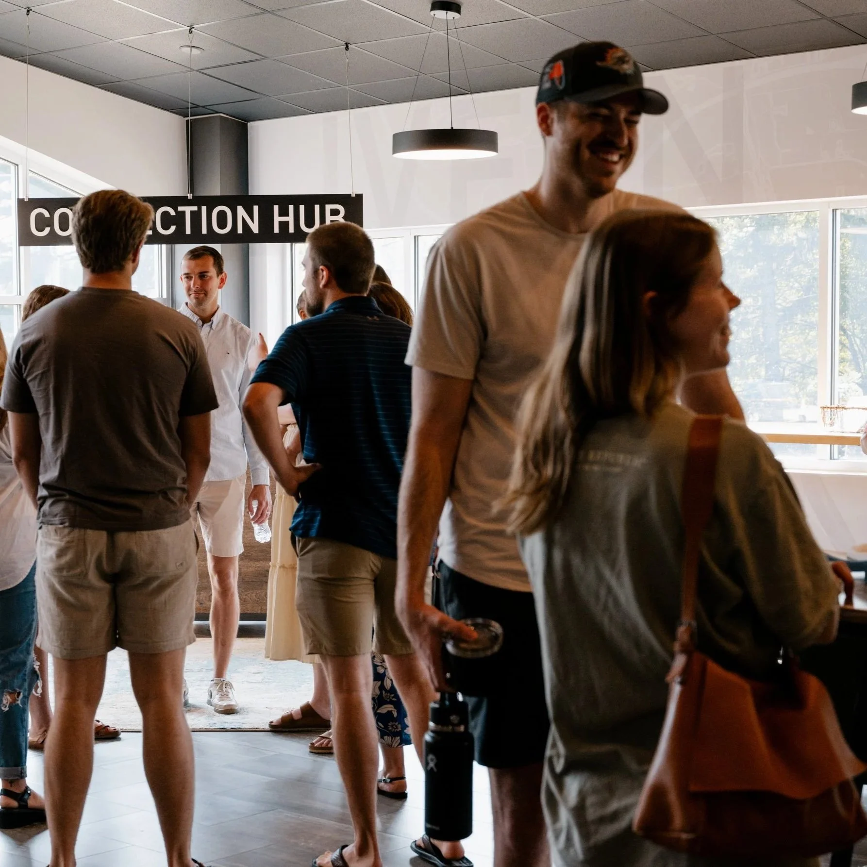 A group of people talking and socializing inside a modern, well-lit room with large windows. There is a sign that reads 'CONNECTION HUB' on the wall.