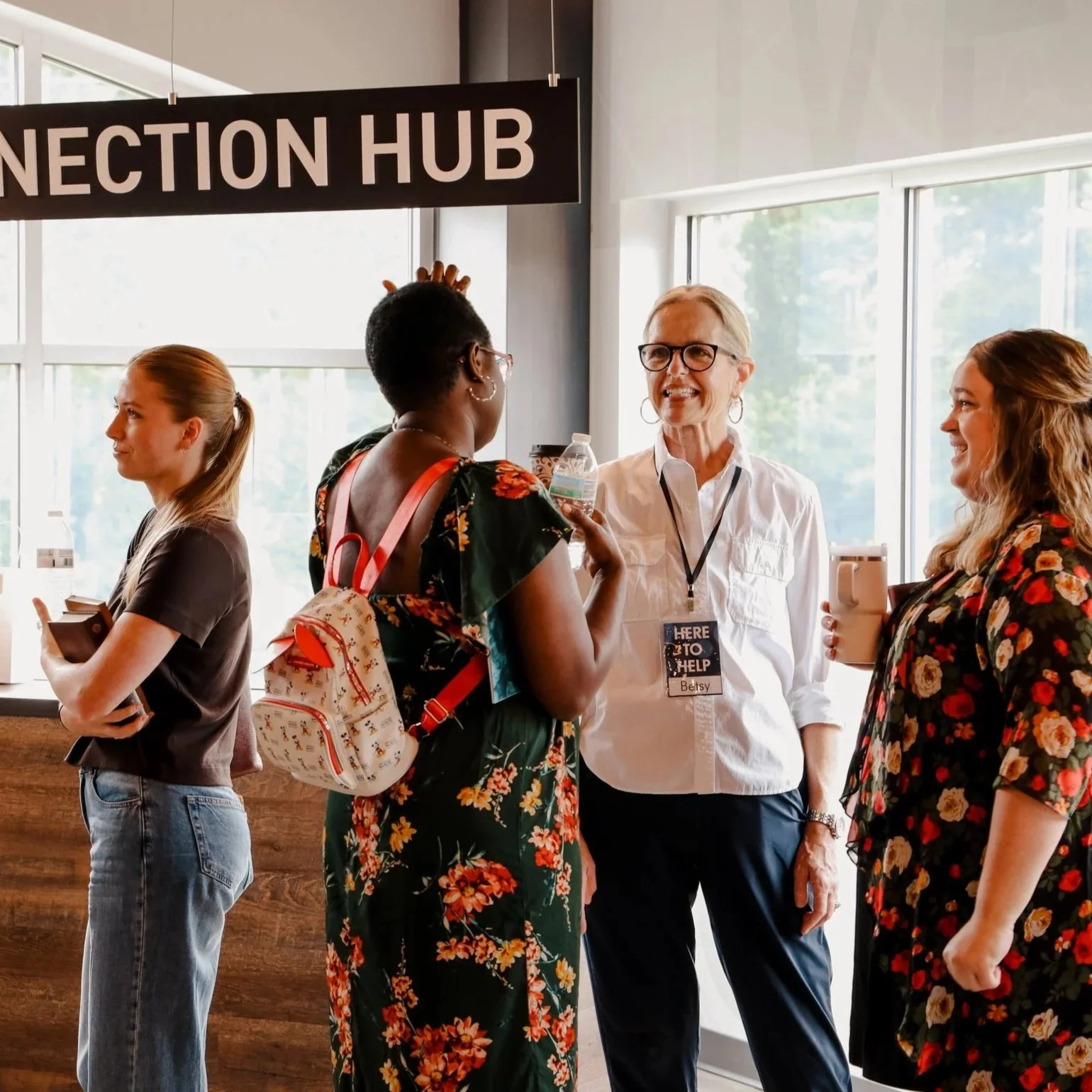 Group of people engaged in conversation at a conference or event near a sign that says 'Connection Hub'. A woman with gray hair and glasses is talking to a woman in a floral dress, while another woman with a ponytail waits nearby holding a coffee mug.