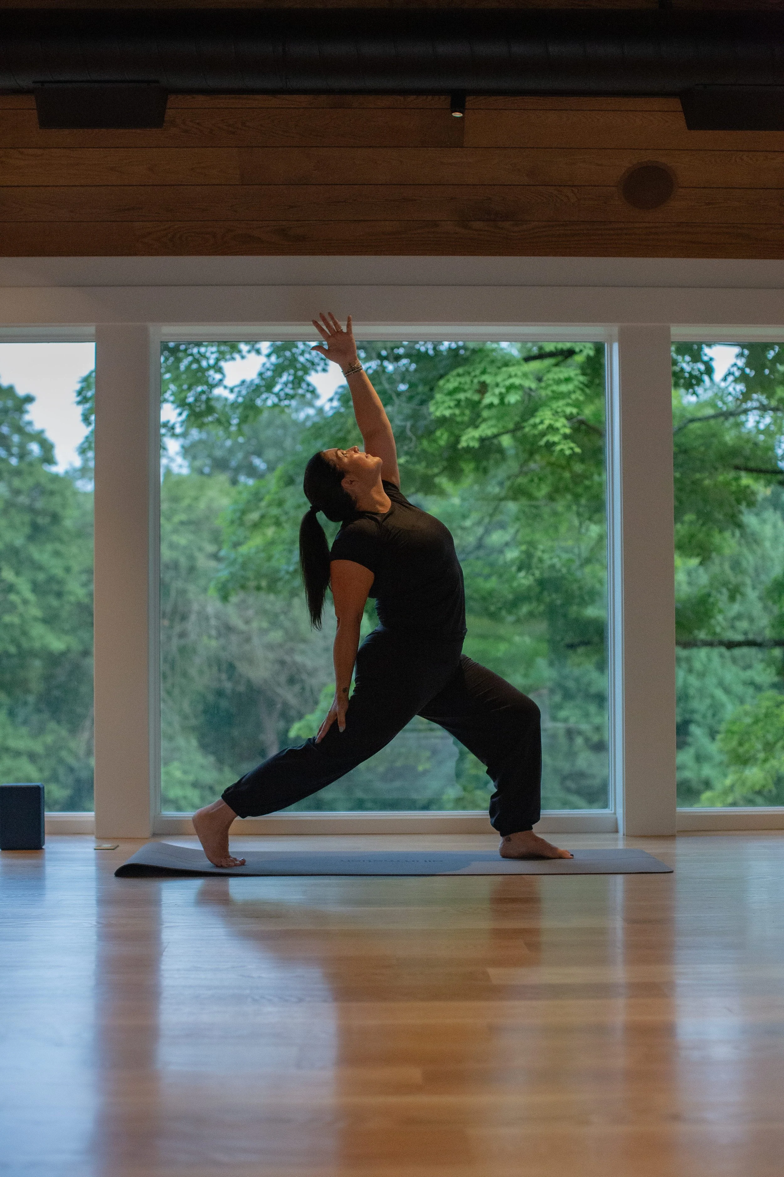 A woman performing a yoga pose indoors with large windows showing green trees outside.