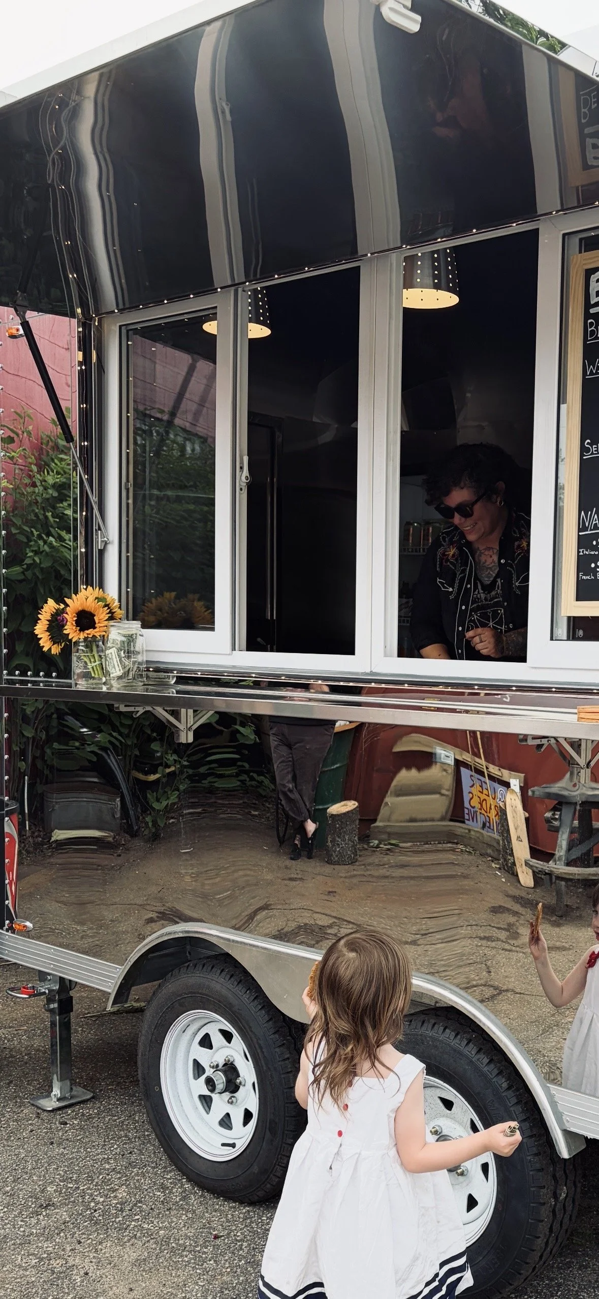A mobile food stand with a black exterior, decorated with fairy lights and sunflowers in jars. The stand has large windows where a woman with short dark hair and sunglasses is serving or preparing food. Two young girls in white dresses with dark stripes are interacting with the stand, one holding a snack.