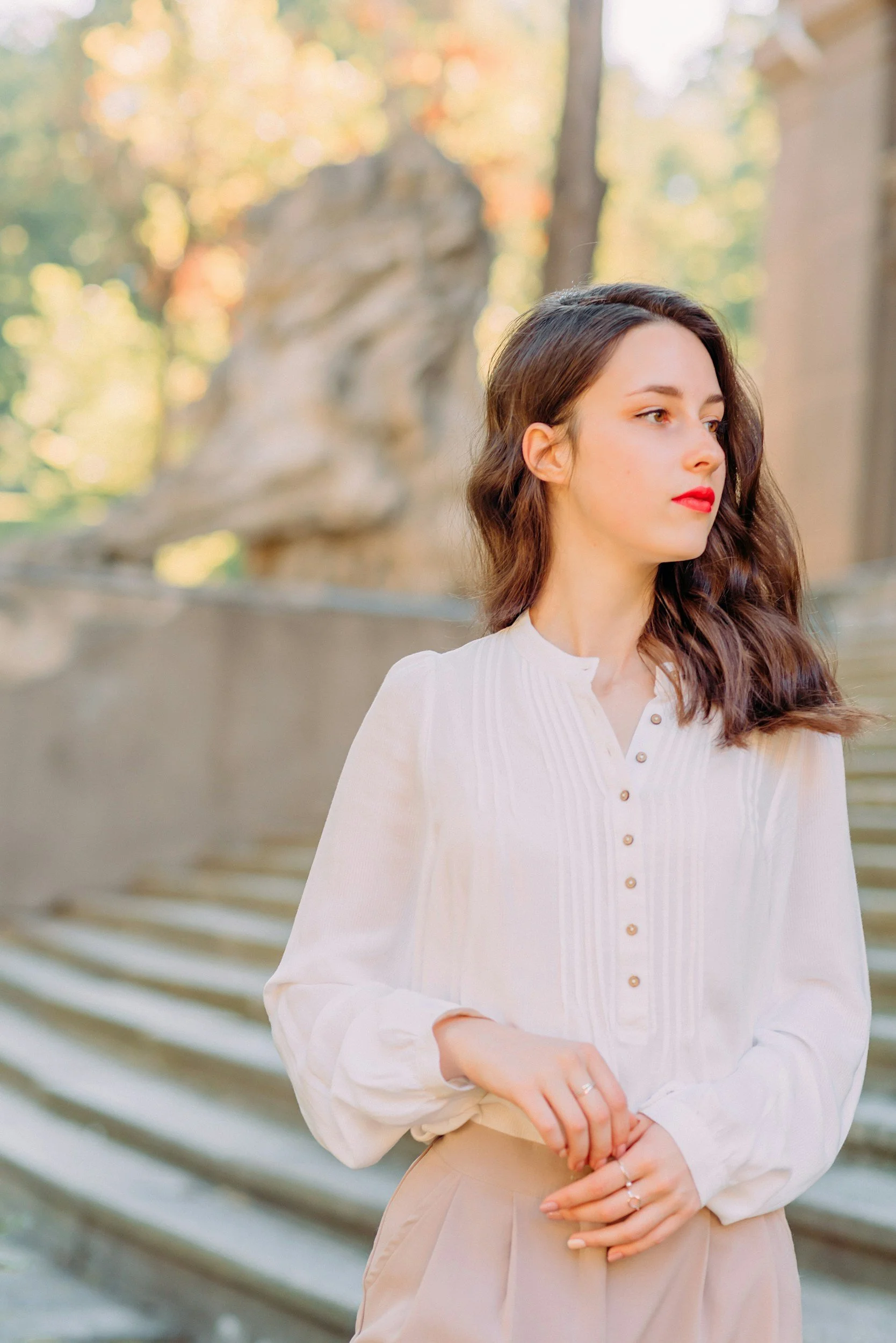 A woman with long brown hair and red lipstick standing outdoors, wearing a white blouse and beige skirt, with a blurred autumnal background of trees and a wooden railing.