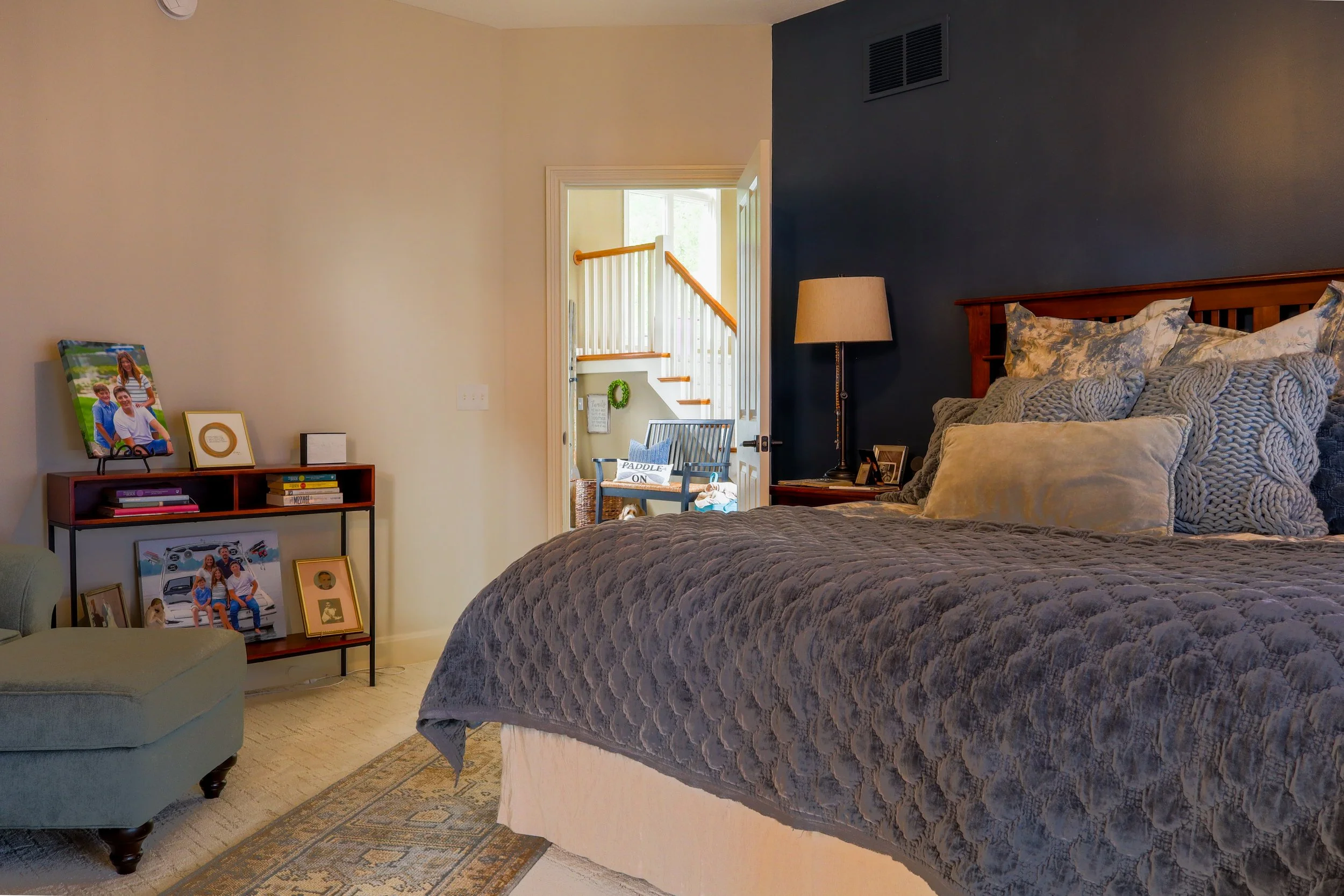 A cozy bedroom with a large bed, dark blue accent wall, wooden headboard, and decorative pillows. Next to the bed is a nightstand with a lamp and picture frames. There is a small seating area visible through the doorway with family photos and a staircase in the background.