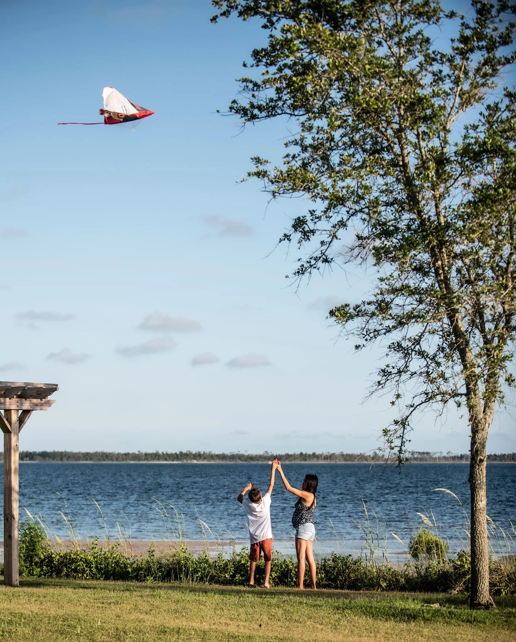 Child and an adult flying a kite beside the water in a public park, illustrating recreation and community connection.