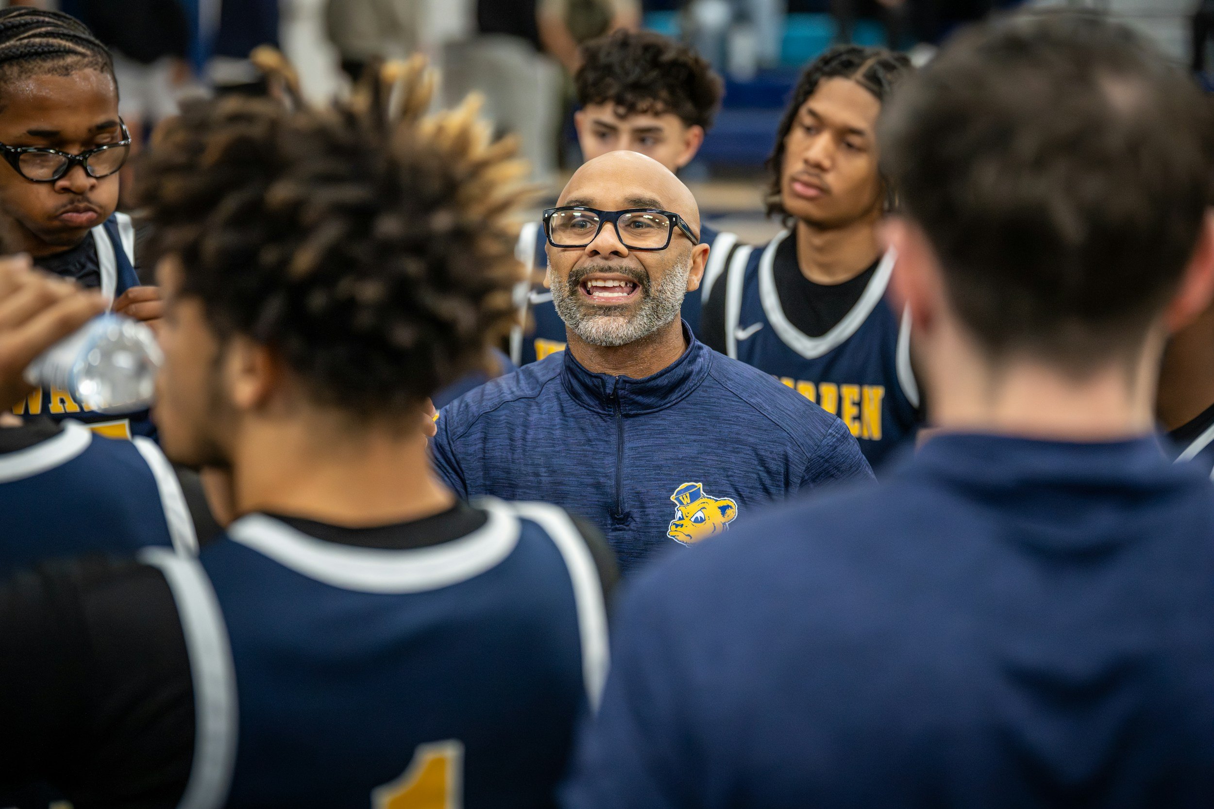 A man with glasses and a beard speaking to a group of young athletes in a team huddle, wearing navy blue sports uniforms with a yellow bulldog logo.