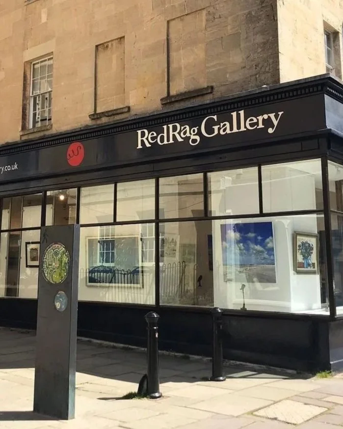 Exterior of Red Rag Gallery with large glass display windows showing artwork inside, a black awning with white lettering, and a sidewalk with a parking meter and black bollards in front.
