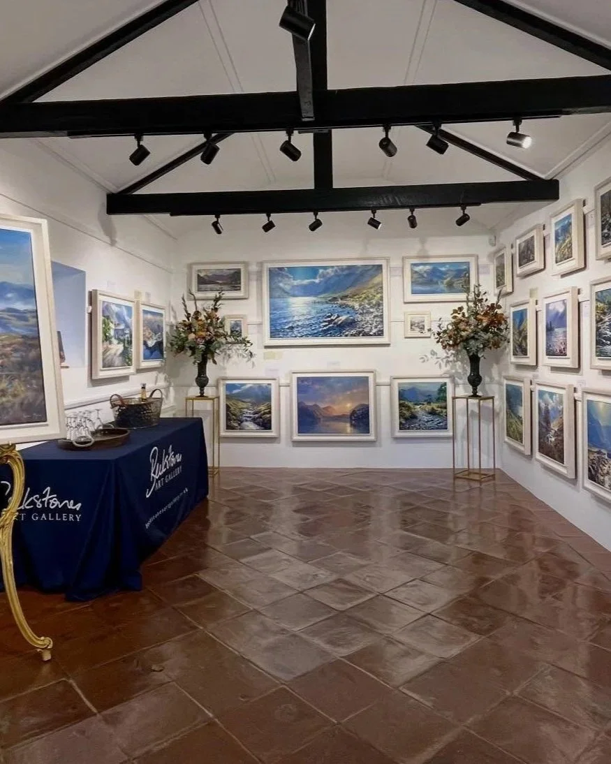 An art gallery wall displaying landscape paintings with two large flower arrangements, a table with a blue cloth, and a brown tiled floor.
