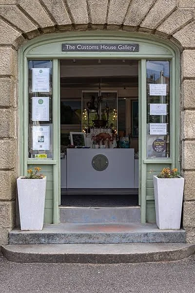 The entrance to The Customs House Gallery features a green wooden door frame with a rounded top, two white planters with small plants on either side, and a sign above the door. Inside, a white reception desk and artwork are visible.