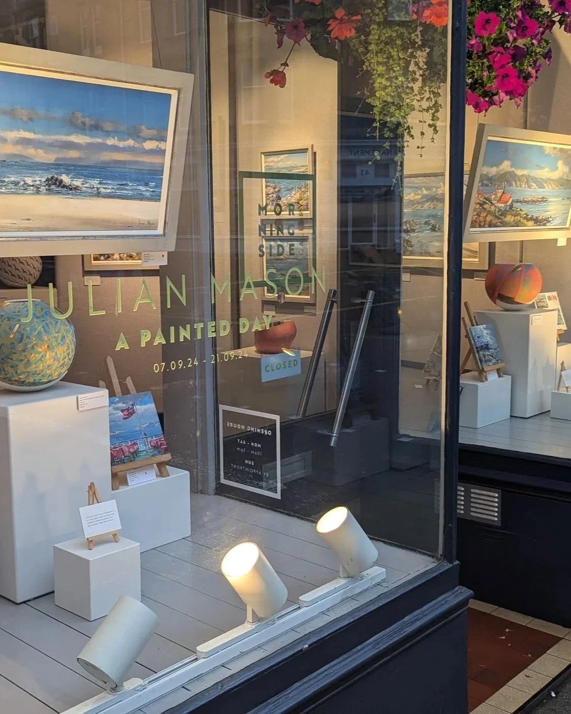 Gallery window display featuring landscape paintings of beaches and coastlines, with hanging flower arrangements and illuminated art pieces inside.
