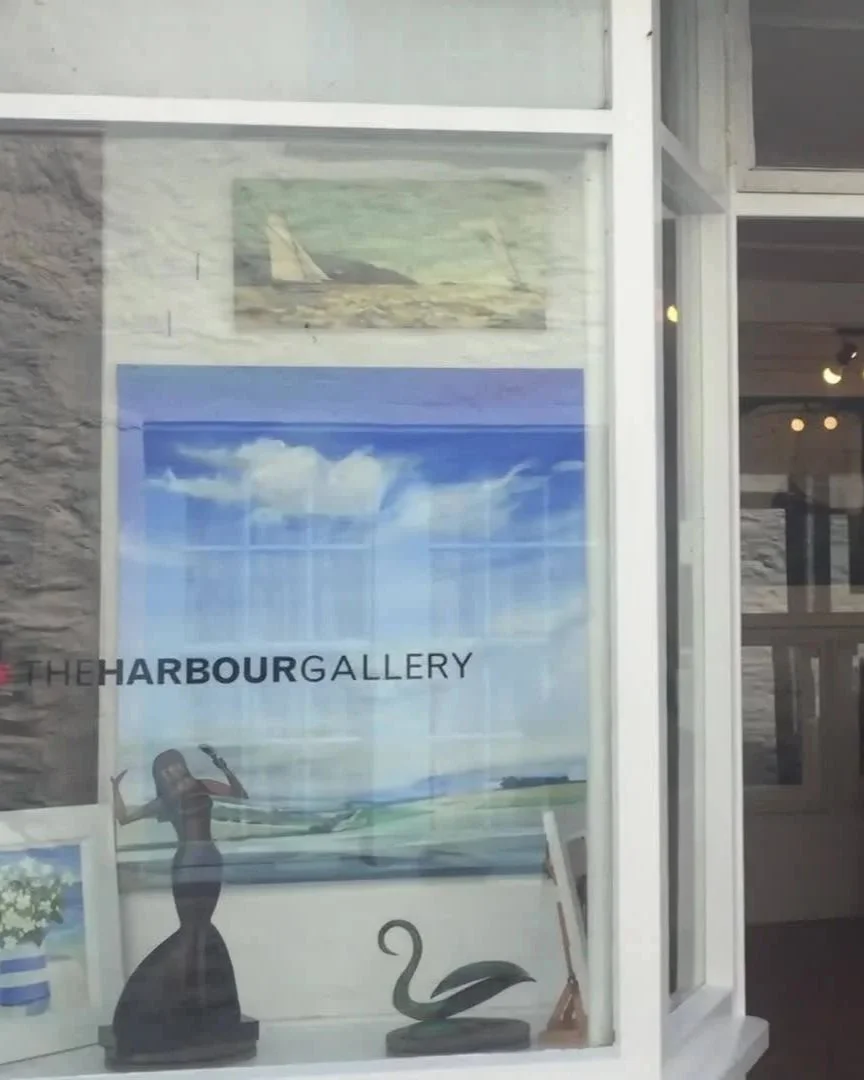Window display at The Harbour Gallery showcasing artworks and decorative items, including paintings, a sculpture of a woman, a swan figure, and a small framed print.