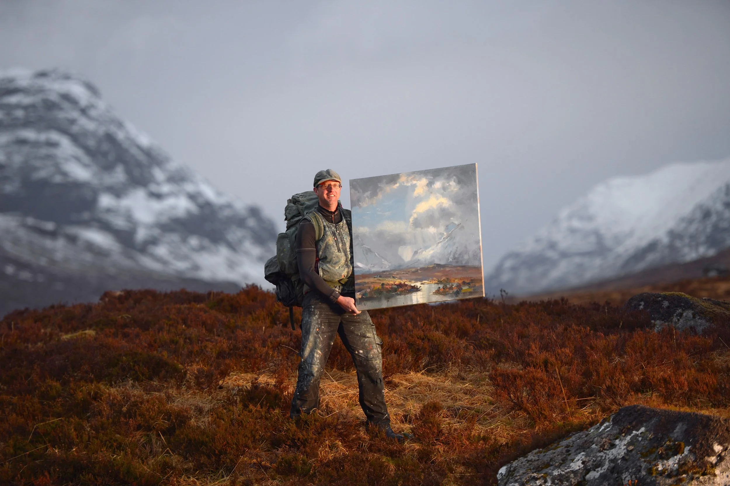 A man stands outdoors on a mountain landscape holding a painting of a sky with clouds, with mountains and cloudy sky in the background.