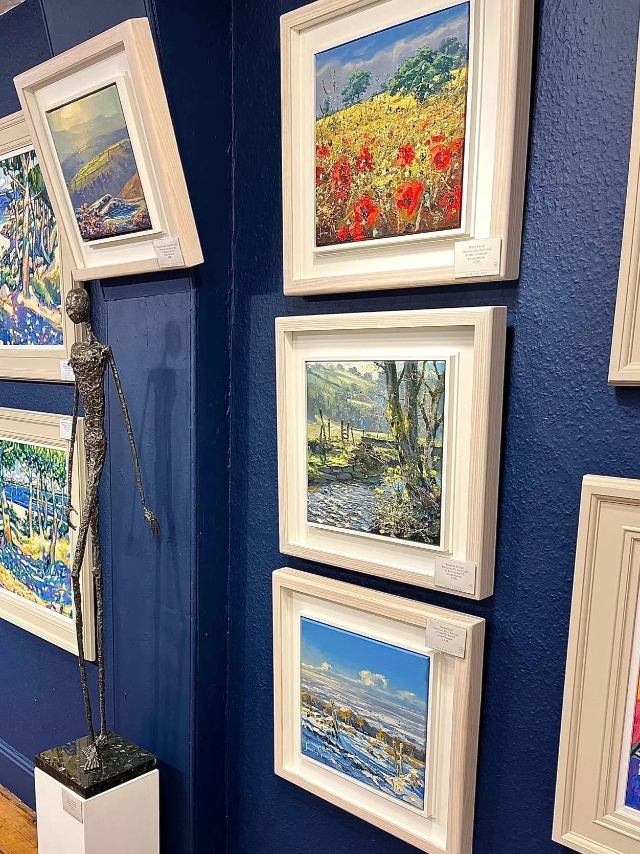 A wall display of landscape paintings in an art gallery, with one sculpture of a slender, elongated human figure placed on a pedestal.