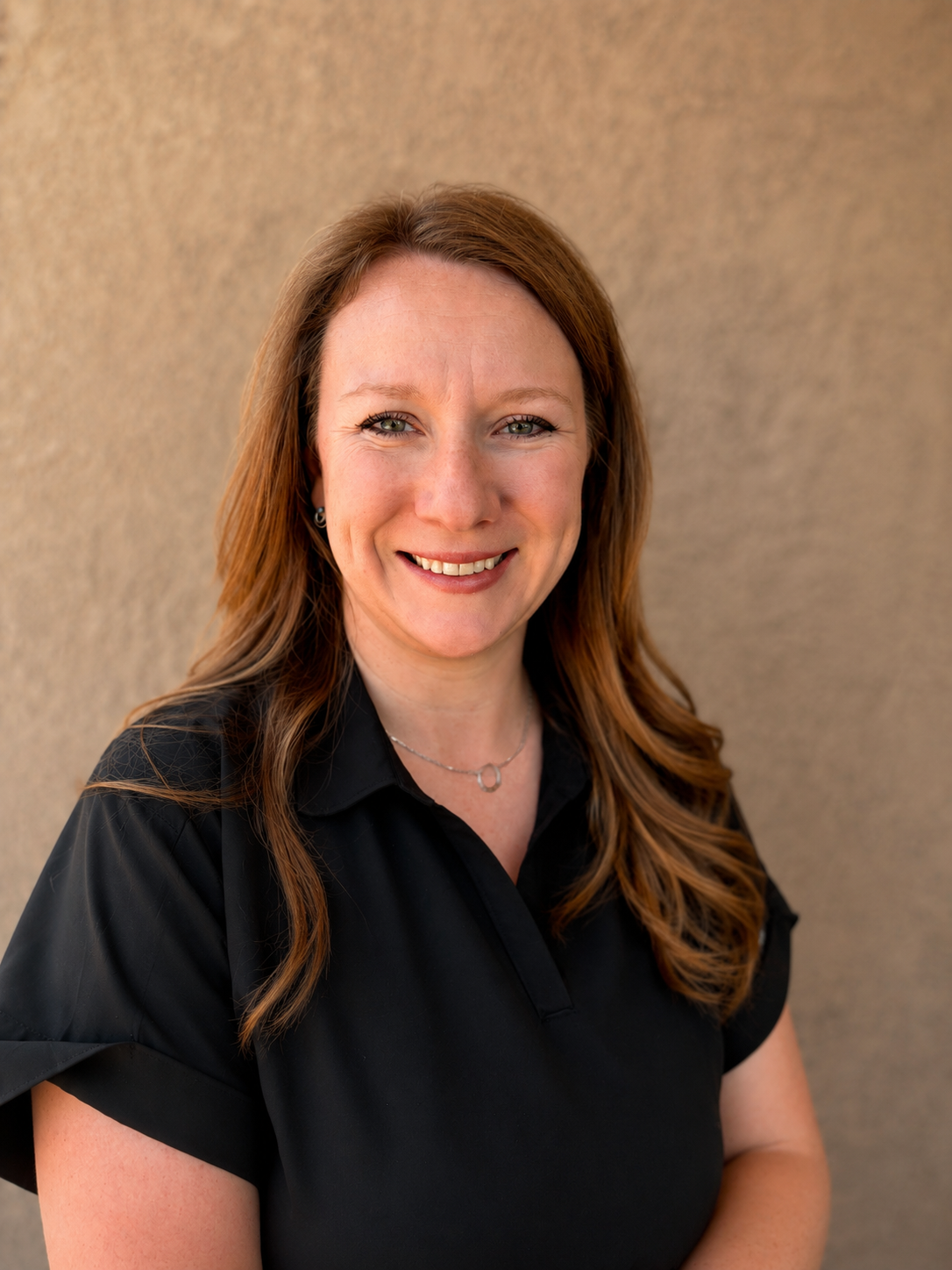 Headshot of a woman with shoulder-length reddish-brown hair smiling, wearing a black shirt and a silver necklace, standing against a beige textured background.