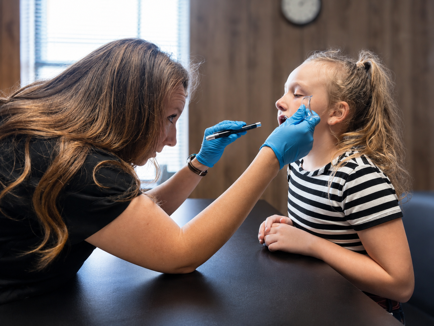 A young girl sitting at a table with her mouth open while a female healthcare professional in blue gloves examines her mouth with a tongue depressor, in a medical office with wood panel walls and a clock in the background.