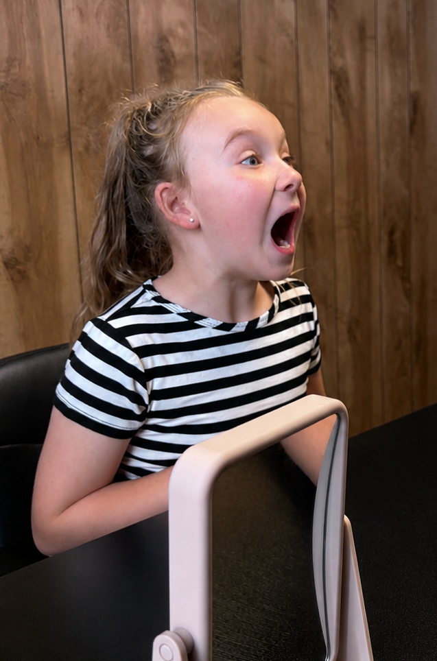 A young girl with curly blonde hair in a striped black and white shirt sitting at a table with her mouth open in surprise or excitement, in front of a wooden wall.